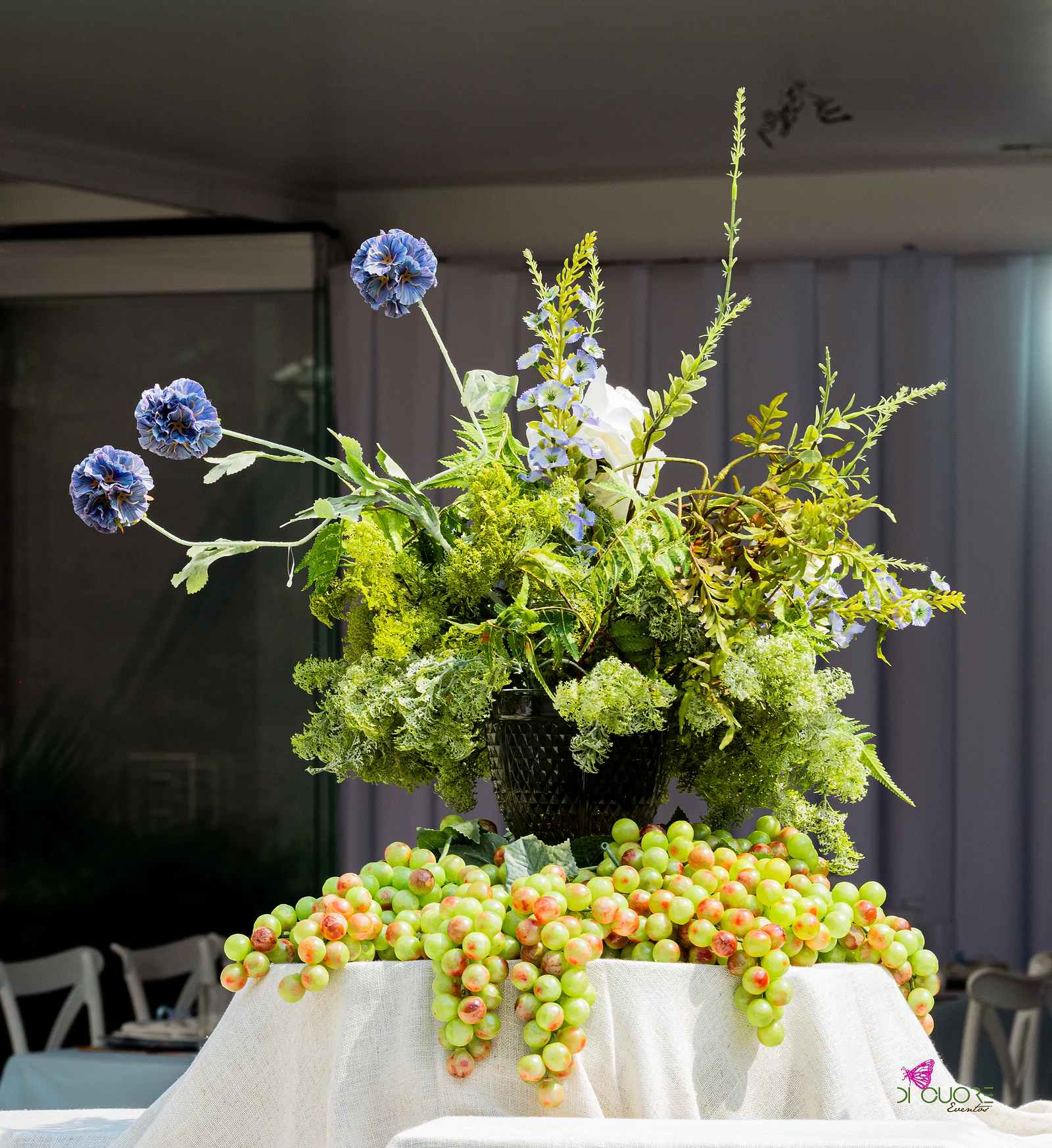 Arranjo de fetos, rosas brancas e flores azuis em bola, sobre cachos de uvas verdes e toalha de linho