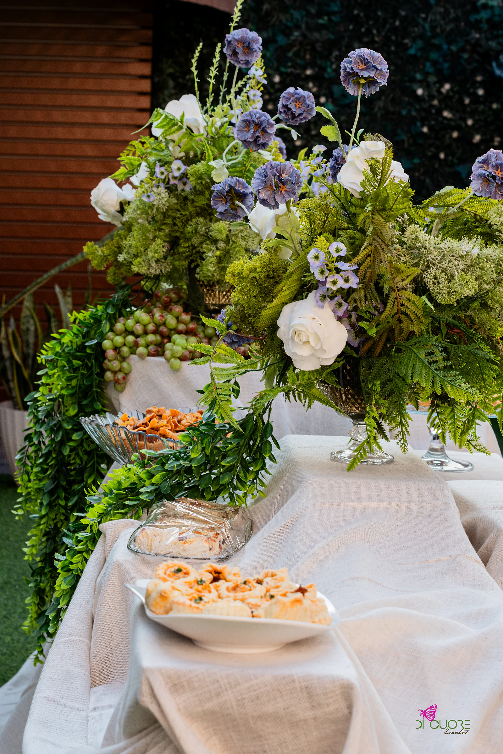 Mesa de buffet com toalha de linho, arranjos de fetos e rosas brancas, uvas e petiscos em travessas