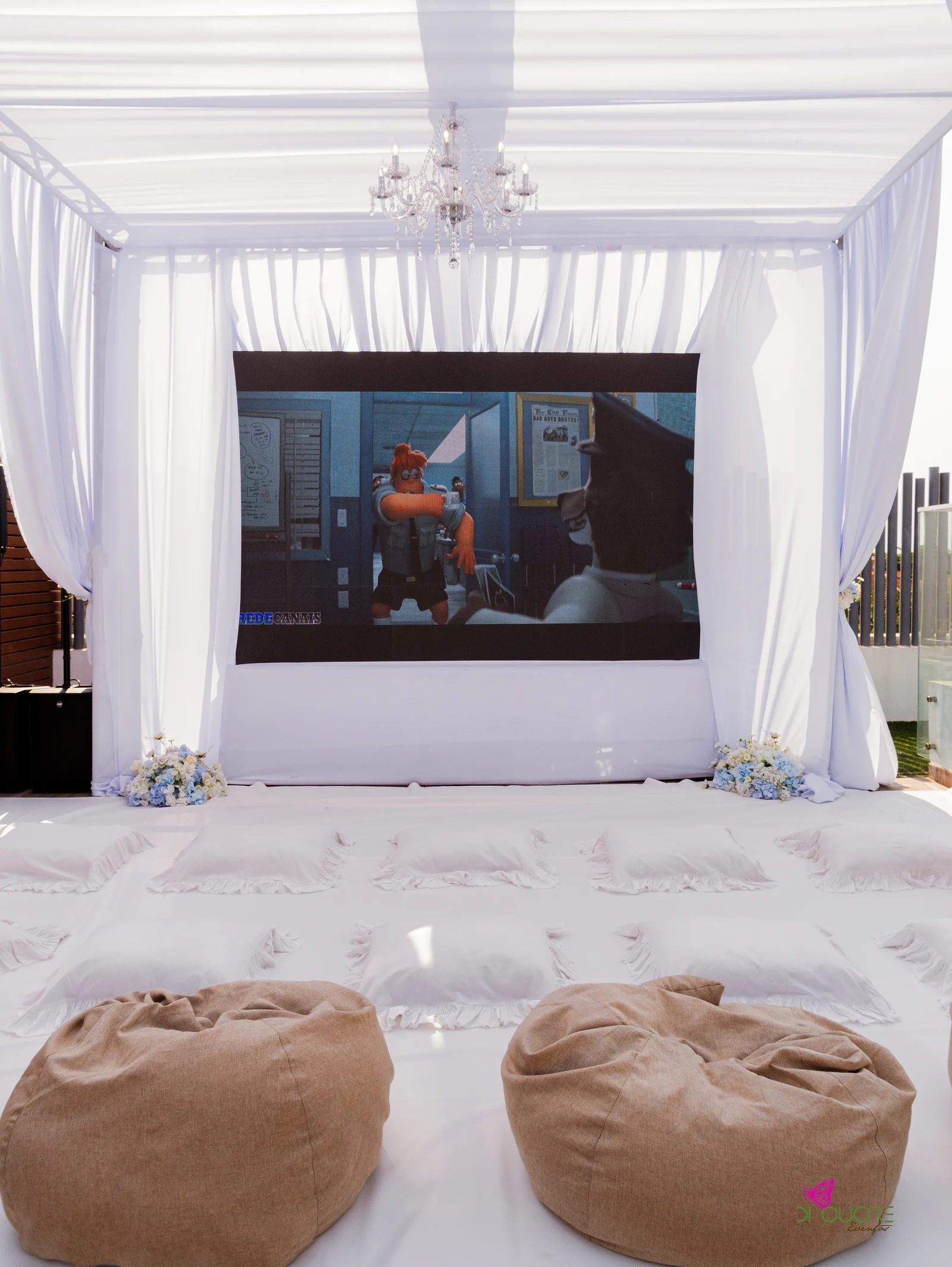 Cantinho de cinema sob um dossel de cortinas brancas, com candeeiro de cristal, ecrã, almofadas brancas e dois puffs cor de areia