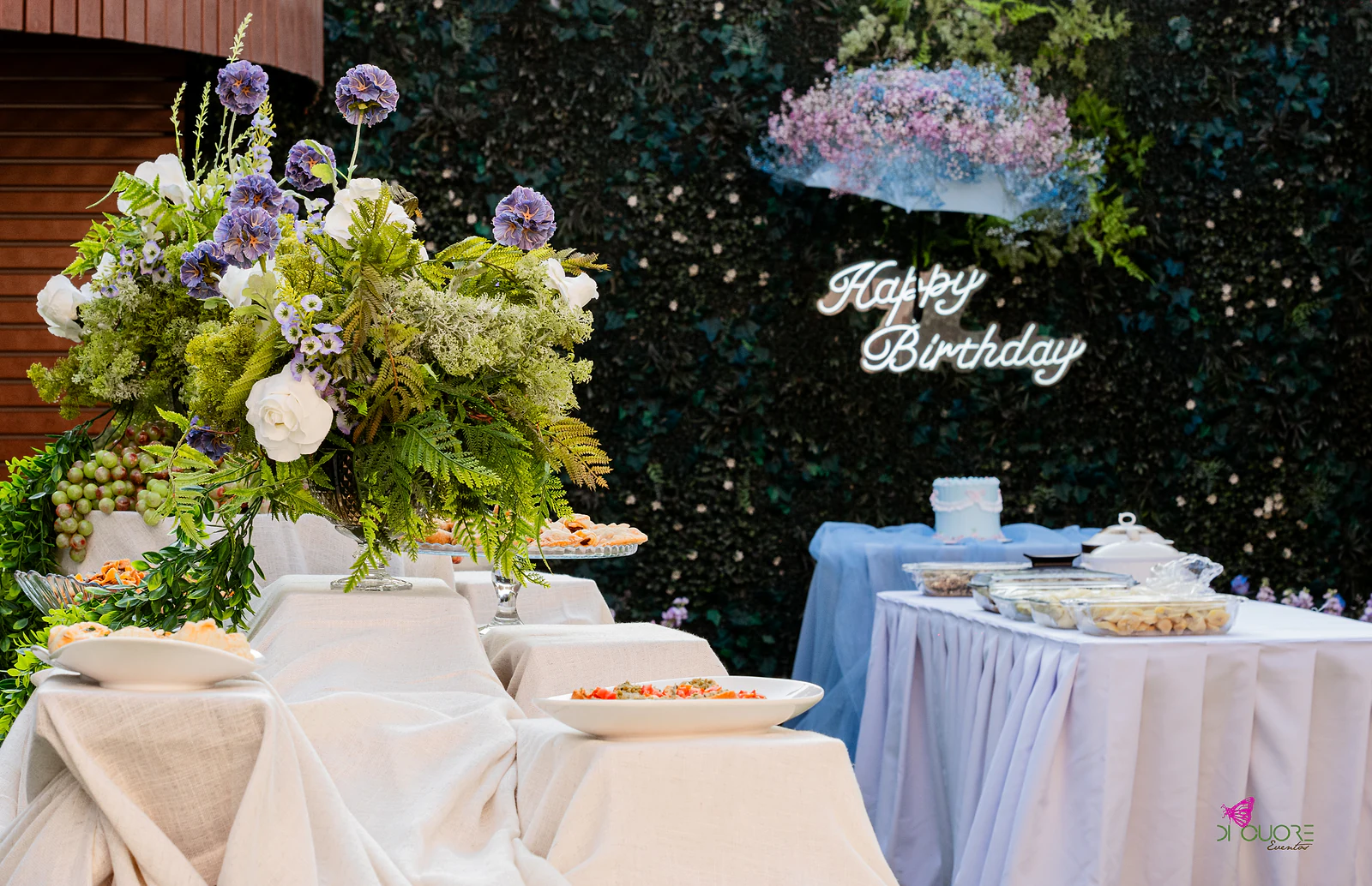 Buffet com toalhas de linho a vários níveis, grandes arranjos de fetos, rosas brancas e flores azuis, e ao fundo o letreiro Happy Birthday