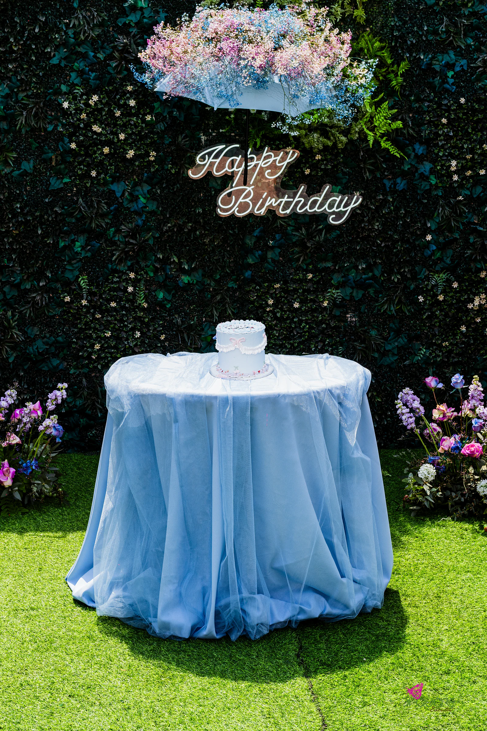 Mesa redonda com tule azul-claro e um bolo, diante de um muro de folhagem com o letreiro Happy Birthday e um guarda-chuva de gypsophila