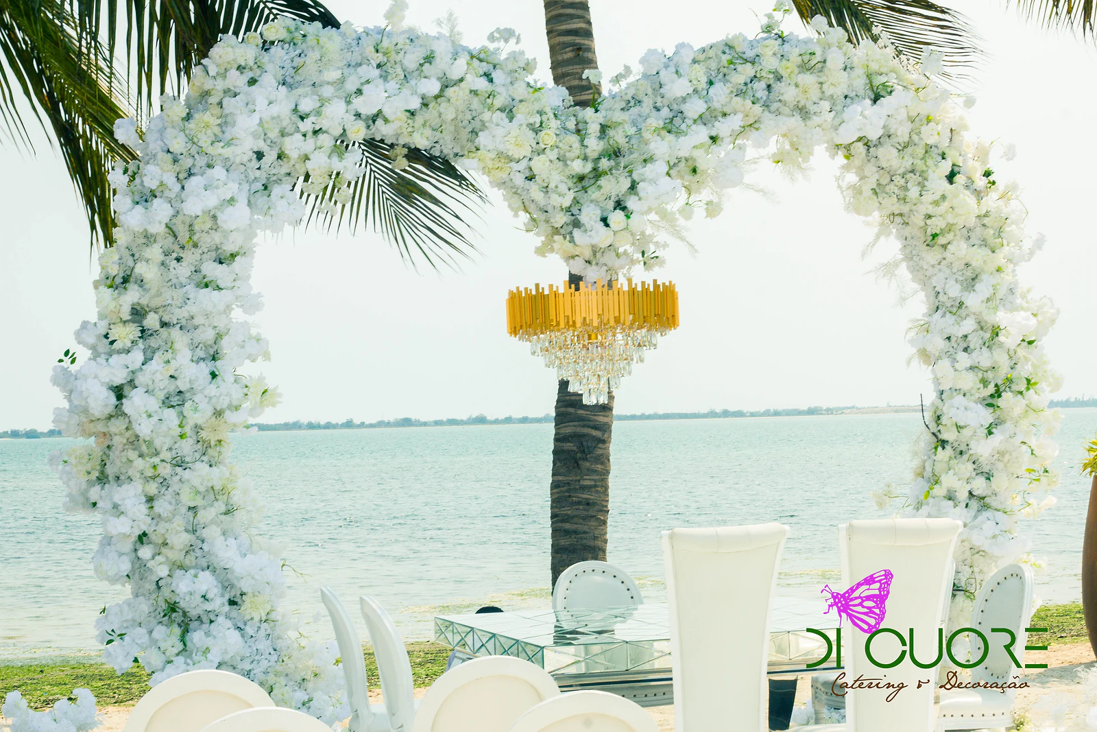 Arco de flores brancas em forma de coração, com um lustre dourado suspenso, a enquadrar o mar e uma palmeira, com cadeiras brancas em primeiro plano