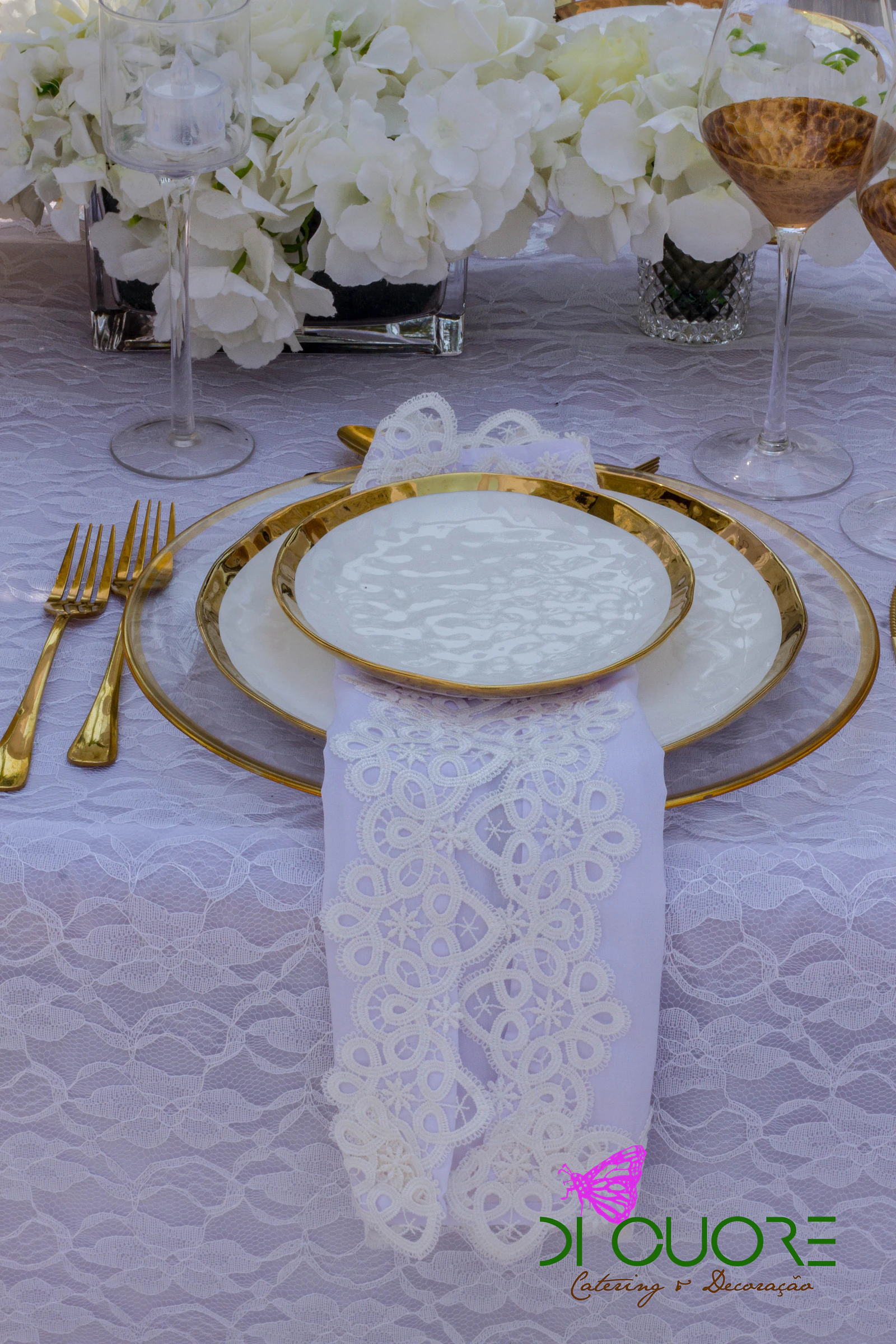 Prato de friso dourado com guardanapo de renda branca sobre uma toalha de renda, com talheres dourados e um copo cor de âmbar