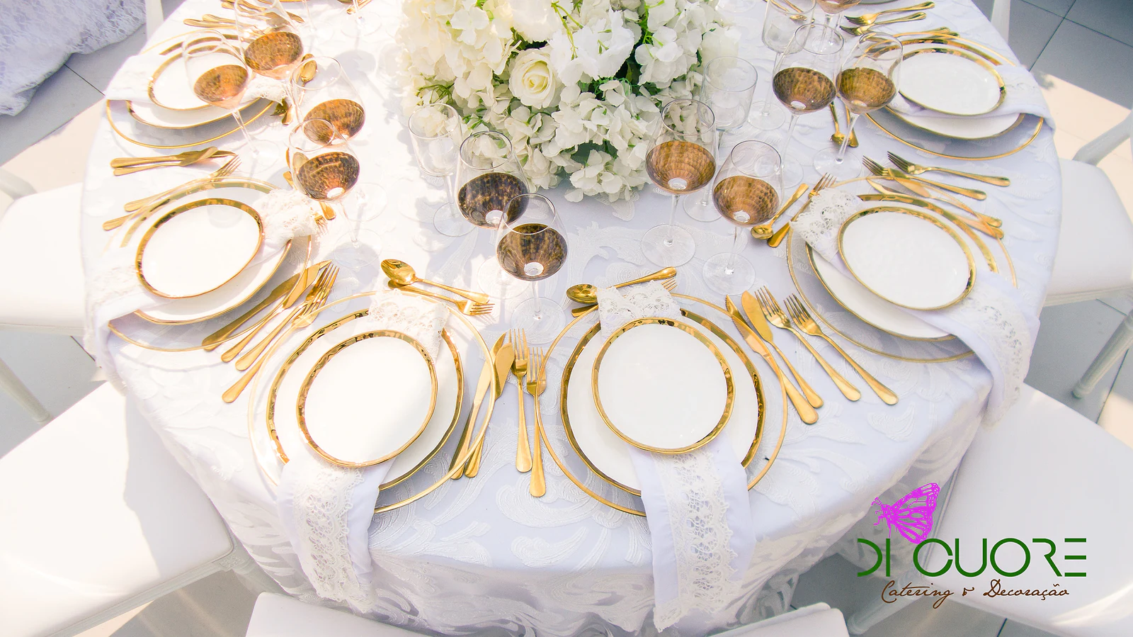 Mesa redonda de toalha branca vista de cima, com pratos de friso dourado, talheres dourados, copos cor de âmbar e um centro de flores brancas