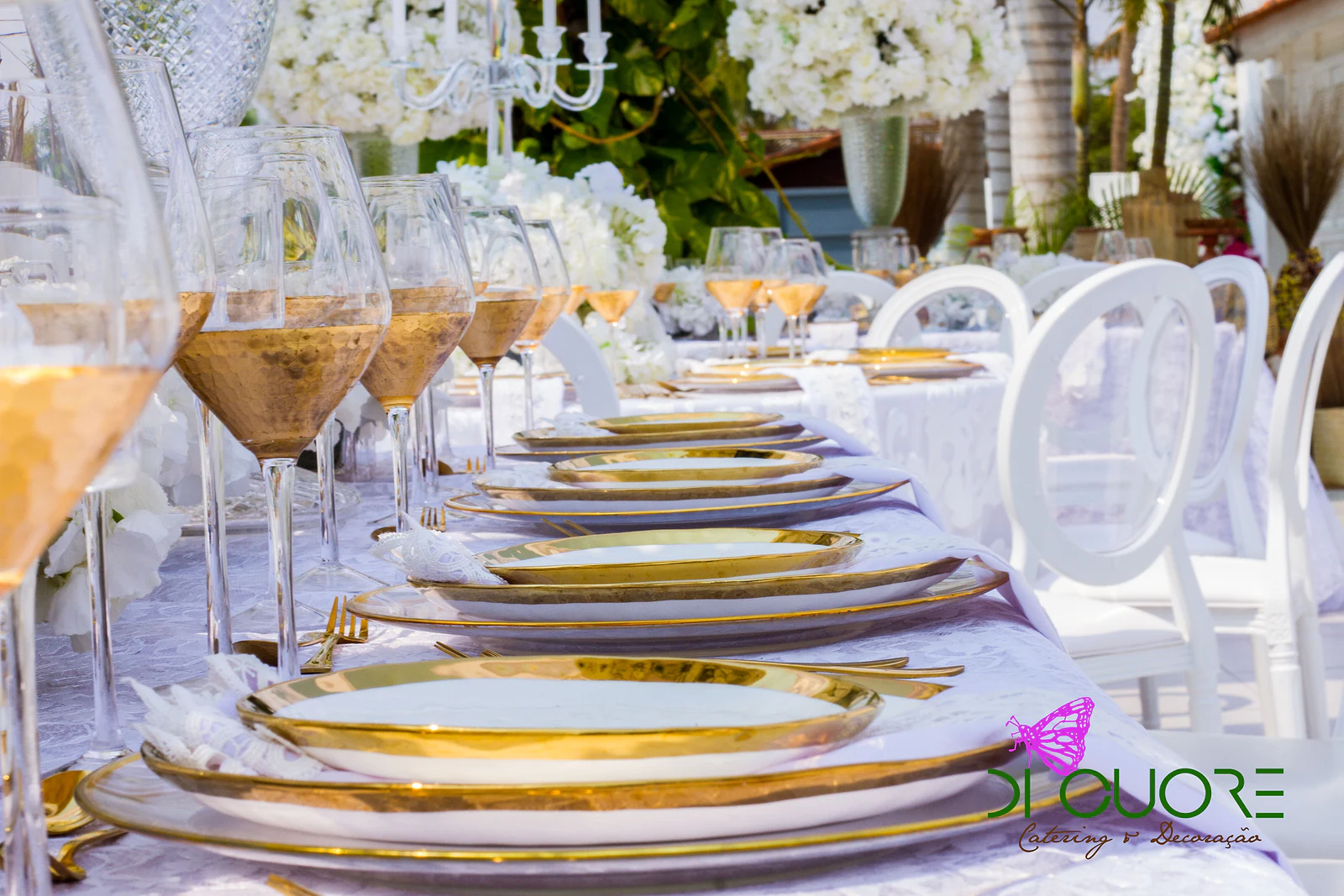 Mesa comprida posta com pratos de friso dourado sobrepostos e copos cor de âmbar, com candelabros e flores brancas ao fundo