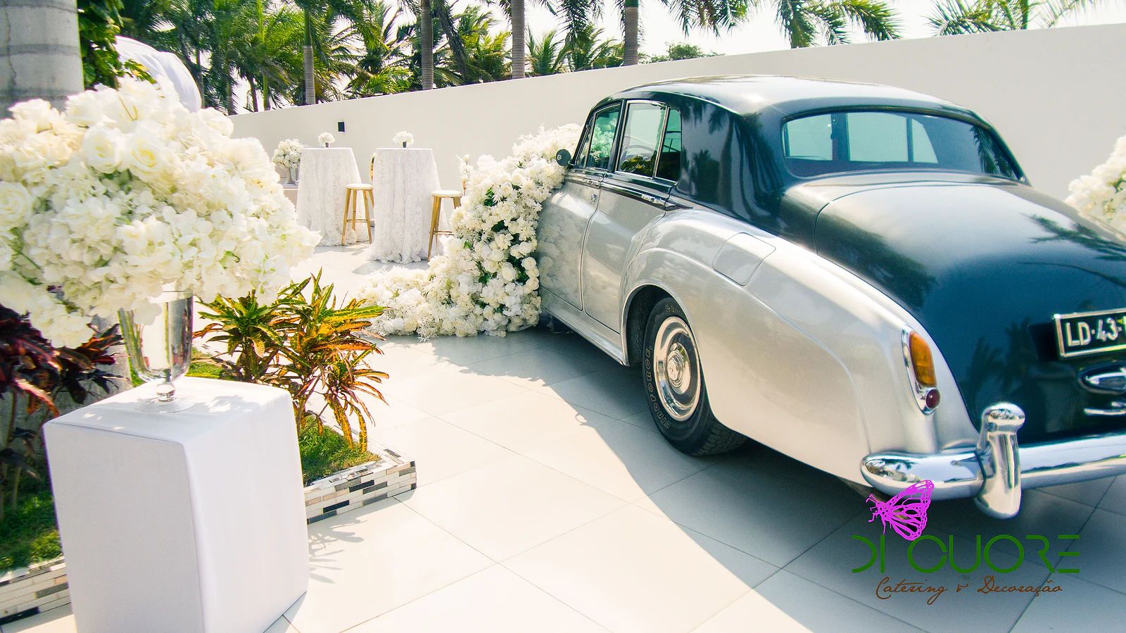 Traseira do carro clássico, com um grande ramo de flores brancas num pedestal branco e mesas altas de renda ao fundo