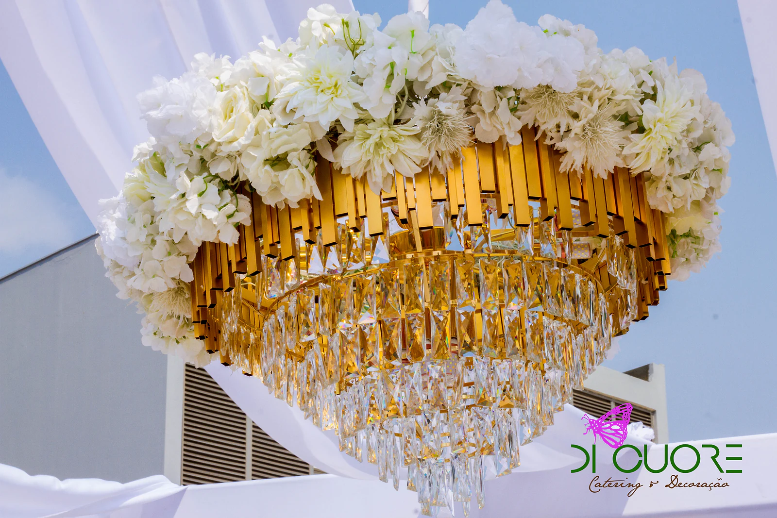 Lustre dourado de cristais coroado de flores brancas, suspenso sob tecidos brancos drapeados, contra o céu azul
