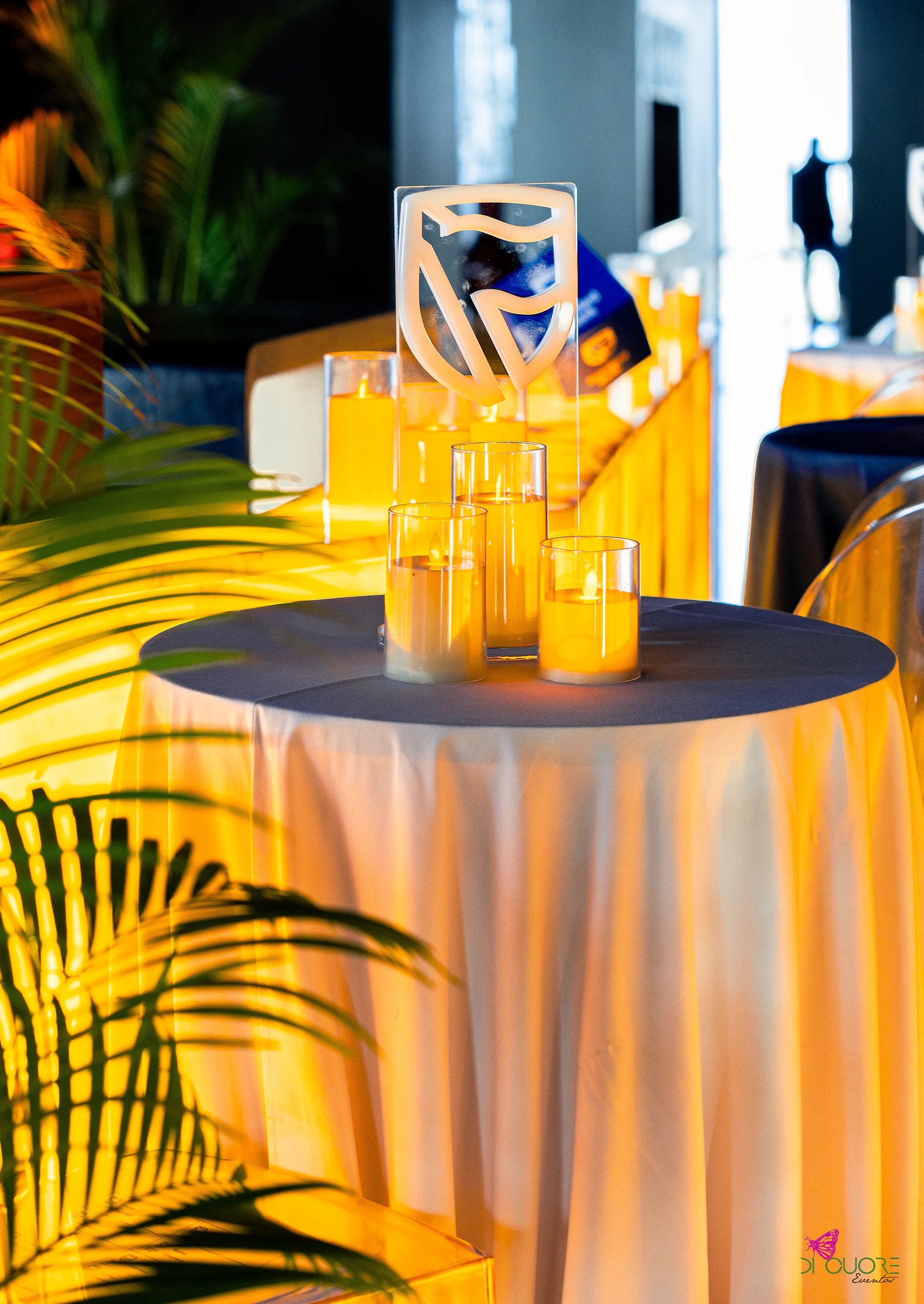 Mesa alta de toalha branca iluminada de âmbar, com velas e o escudo da marca em acrílico, entre folhas de palmeira