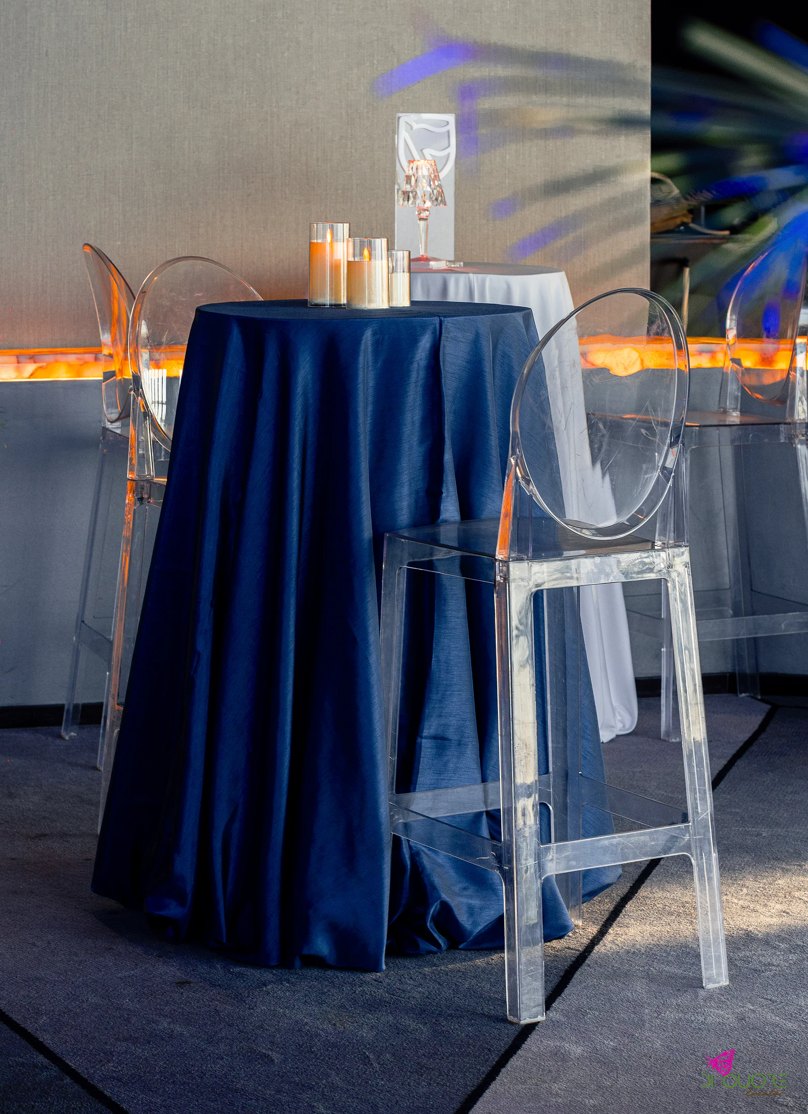 Mesa alta de toalha azul-marinho com velas e escudo em acrílico, ao lado de uma cadeira transparente e do balcão iluminado