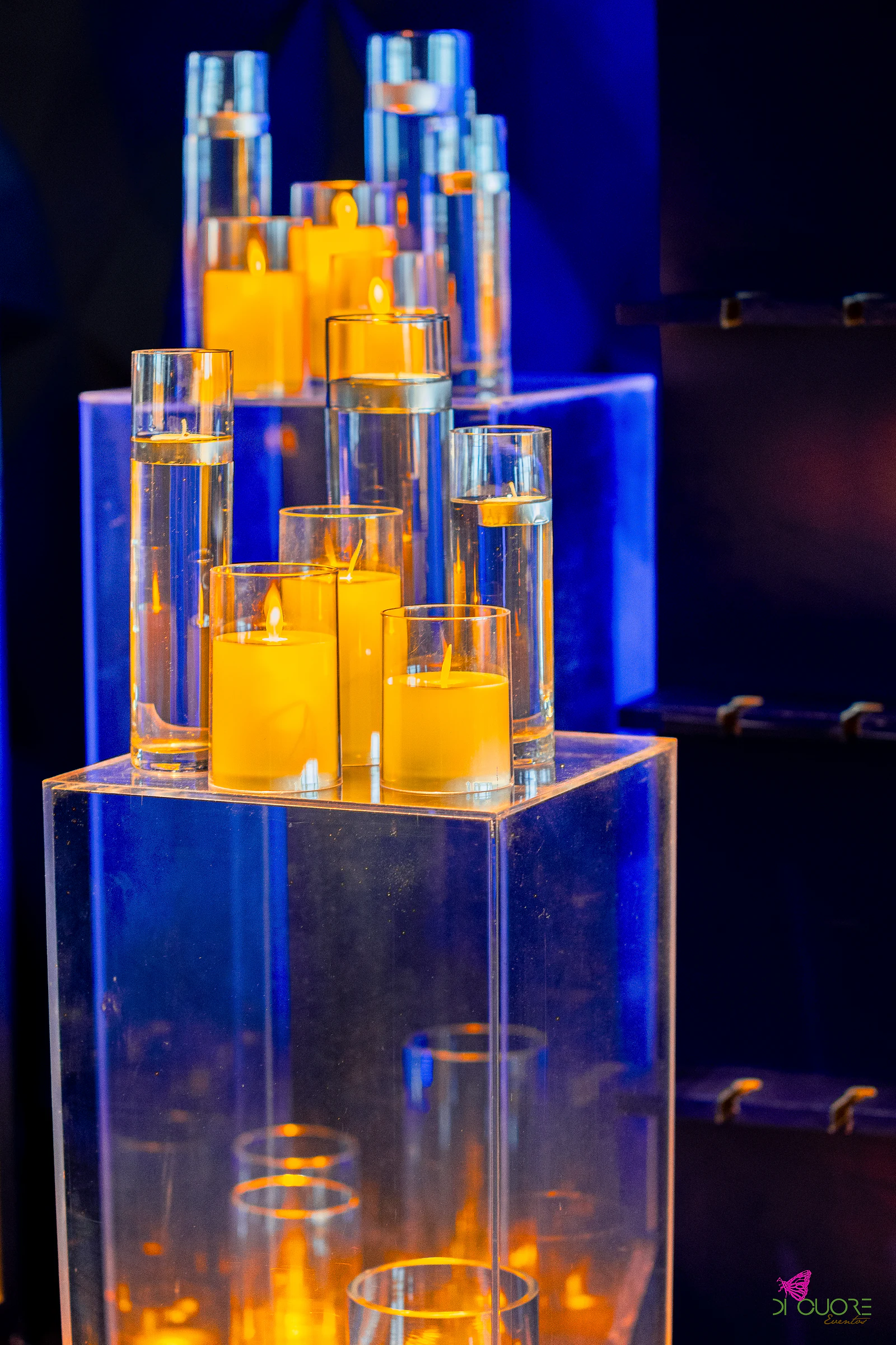 Plintos de acrílico transparente com velas em copos de vidro de várias alturas, sob luz azul