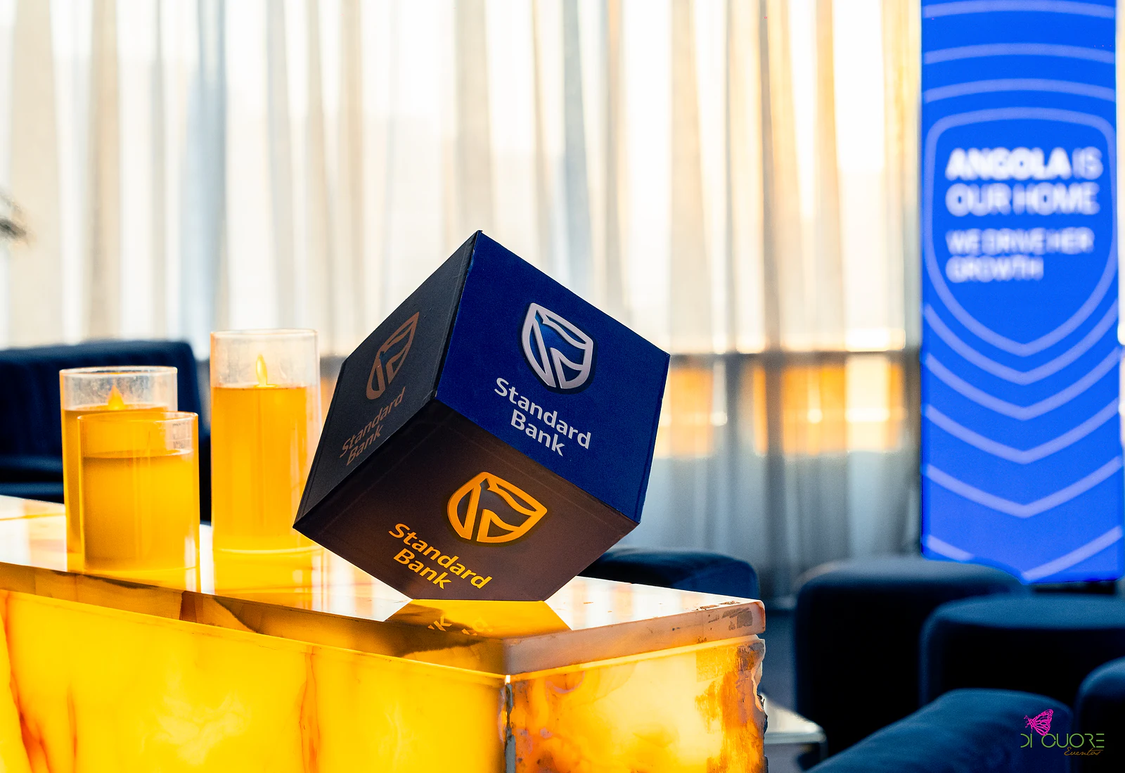 Cubo com o logótipo da marca sobre o balcão de pedra iluminado, ao lado de velas, com sofás azuis e uma cortina branca ao fundo