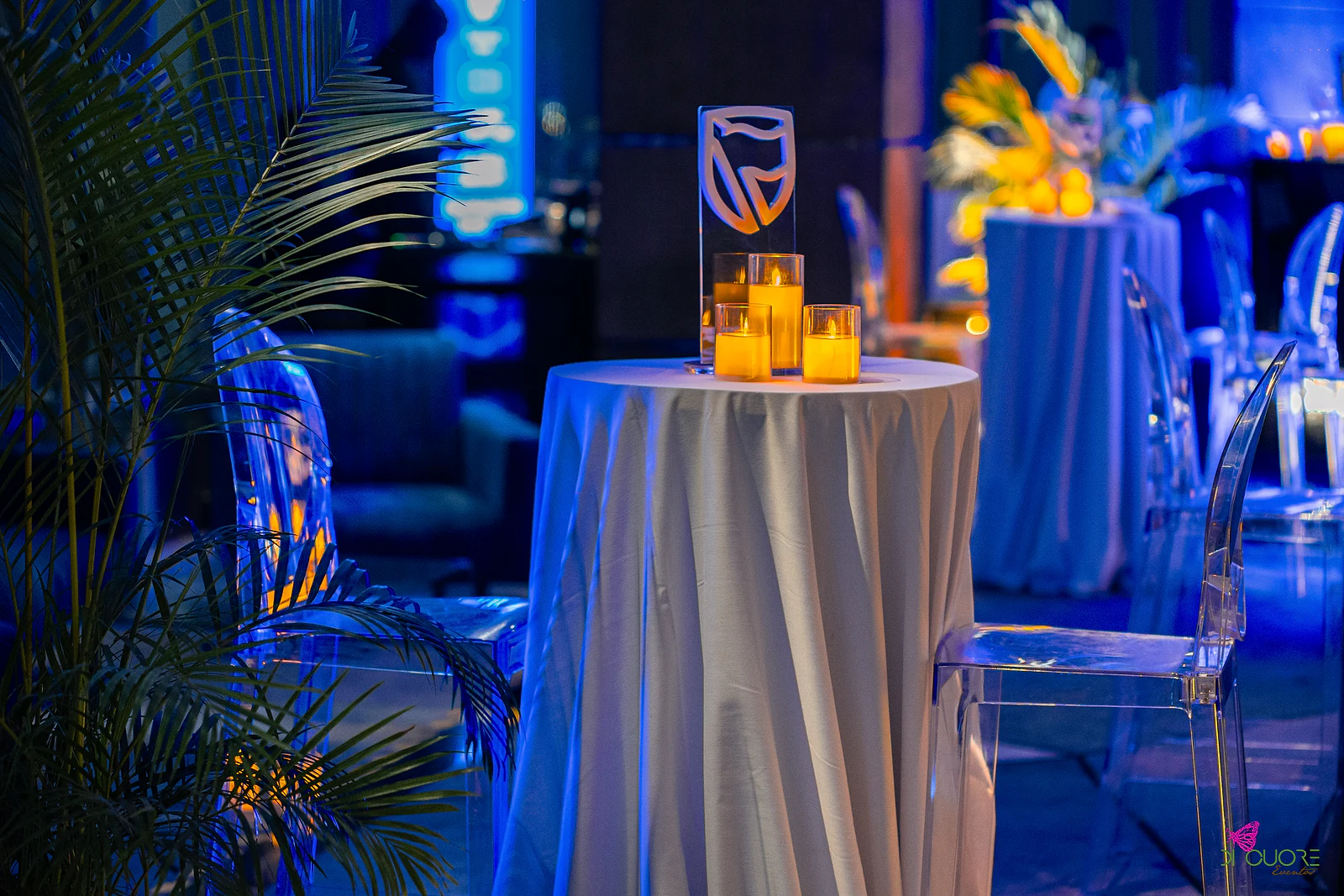 Mesa alta de toalha branca com velas e escudo em acrílico, entre cadeiras transparentes e uma palmeira, em luz azul