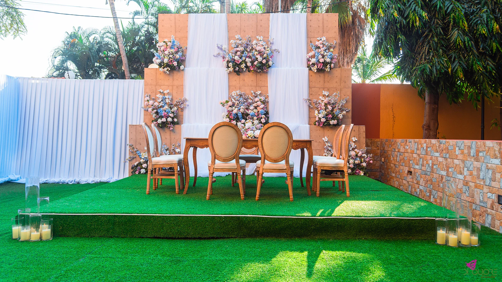 Mesa da cerimónia com cadeiras de madeira de espaldar oval sobre um estrado de relva, diante de um painel com cortinas de tule branco e ramos de flores, e cilindros de vidro com velas nos cantos