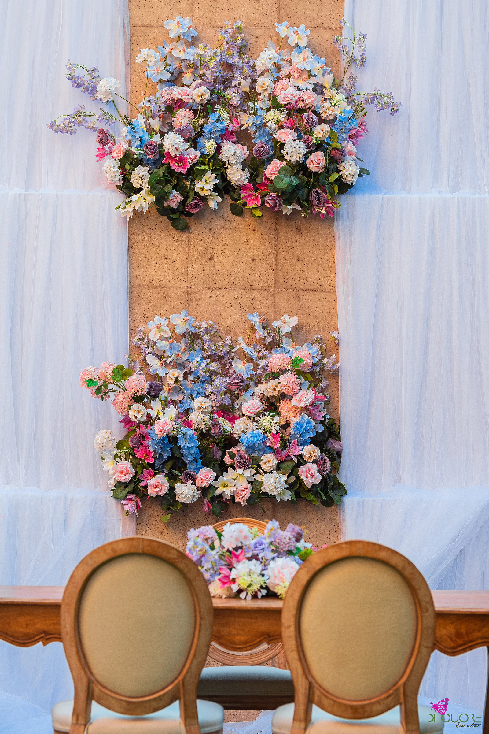Painel de madeira clara com cortinas brancas e dois ramos de flores em rosa e azul, sobre a mesa da cerimónia com cadeiras