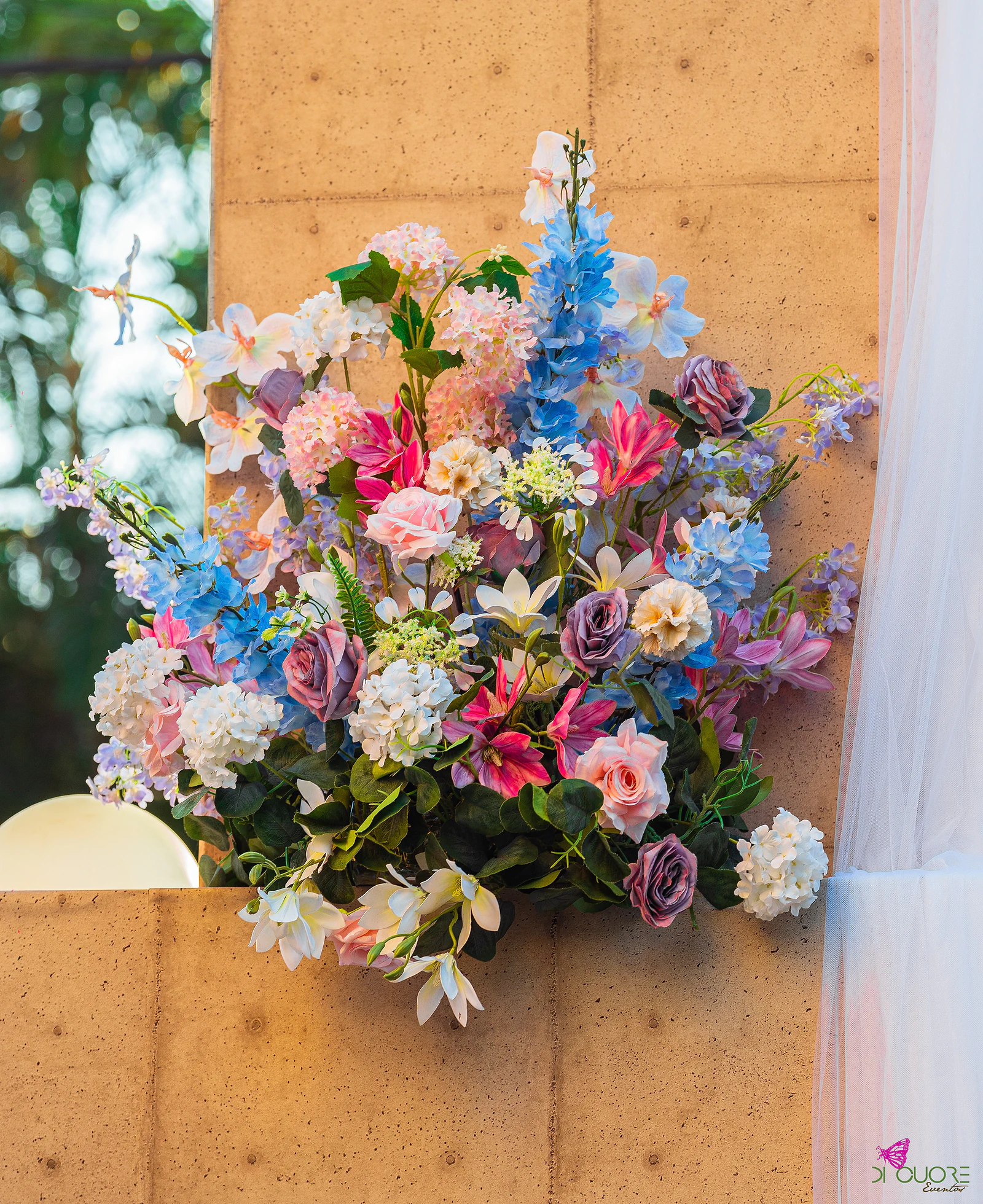 Ramo de flores azuis, cor-de-rosa, brancas e lilás preso a um painel de madeira clara, ao lado de uma cortina de tule branco