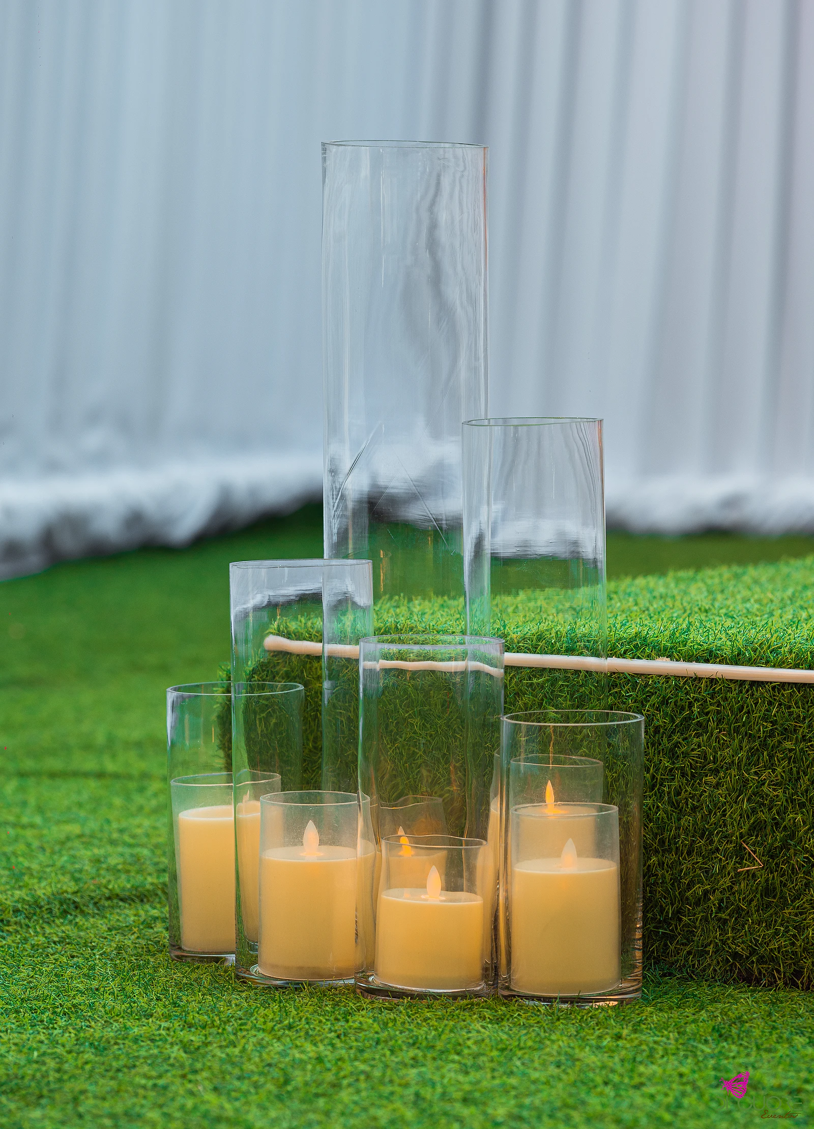 Velas em cilindros de vidro de várias alturas sobre a relva, diante de um tecido branco