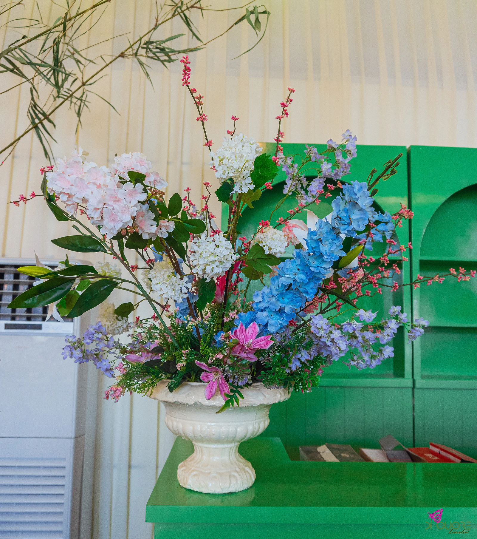 Jarra branca com hortênsias, delfínios azuis e flores cor-de-rosa sobre um bar verde, diante de cortinados claros e ramos de bambu