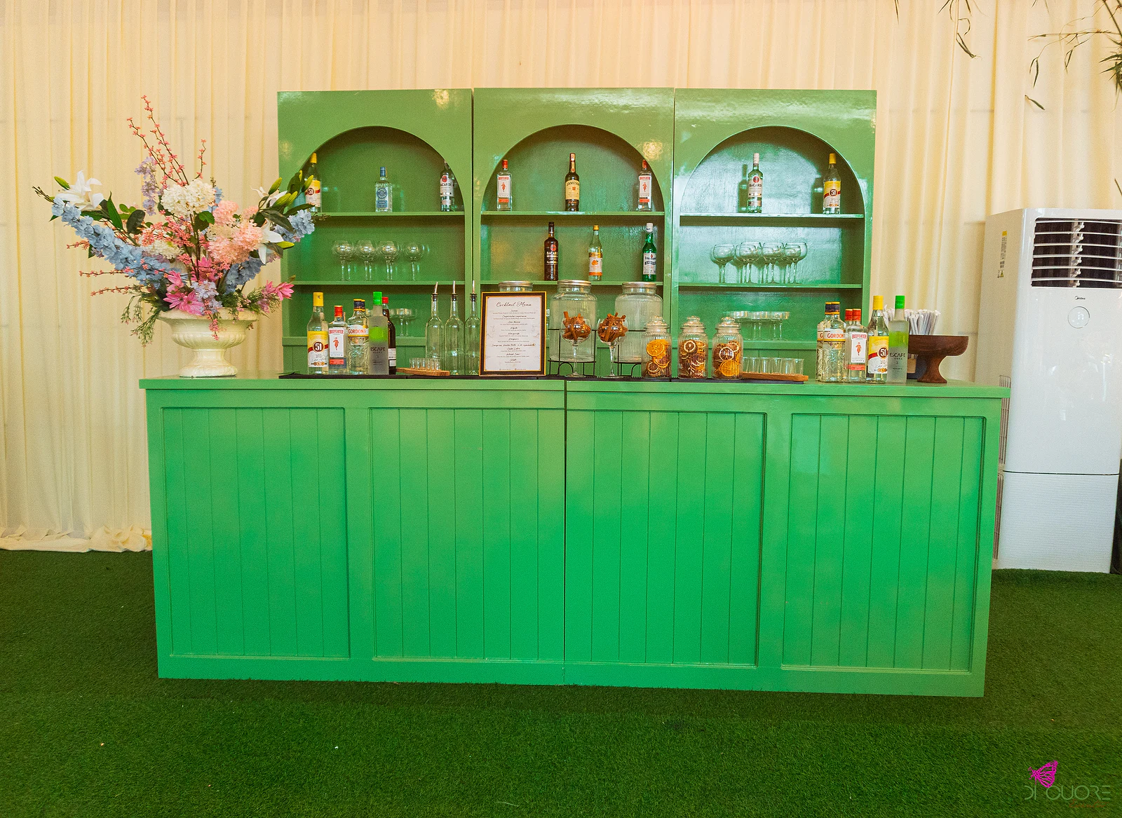 Bar verde com três prateleiras em arco, garrafas, copos e frascos de vidro, e uma jarra branca com flores azuis e cor-de-rosa