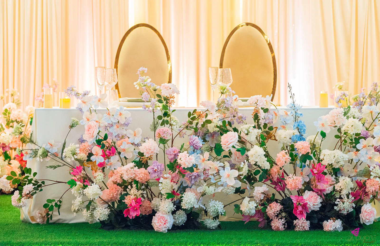 Mesa dos noivos de saia branca coberta de flores cor-de-rosa, brancas e lilás ao nível da relva, com duas cadeiras ovais de moldura dourada diante dos cortinados