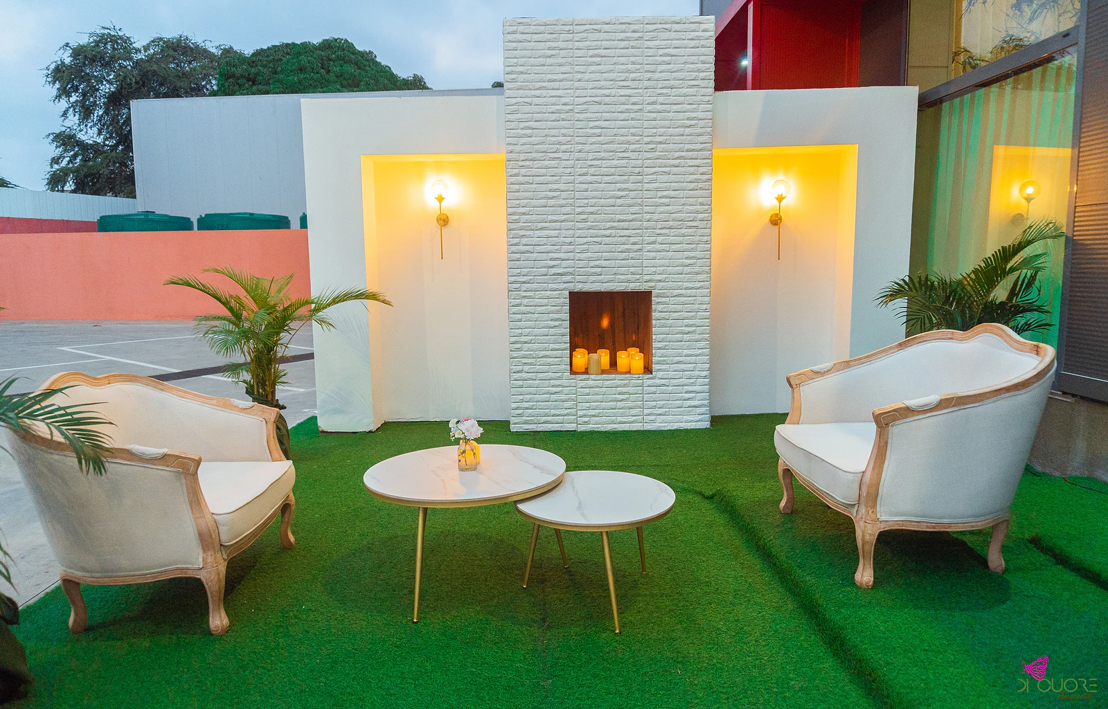 Lounge exterior com dois sofás de madeira clara e estofo creme, duas mesas baixas de tampo em mármore, uma lareira de tijolo branco com velas e candeeiros de parede acesos, sobre relva