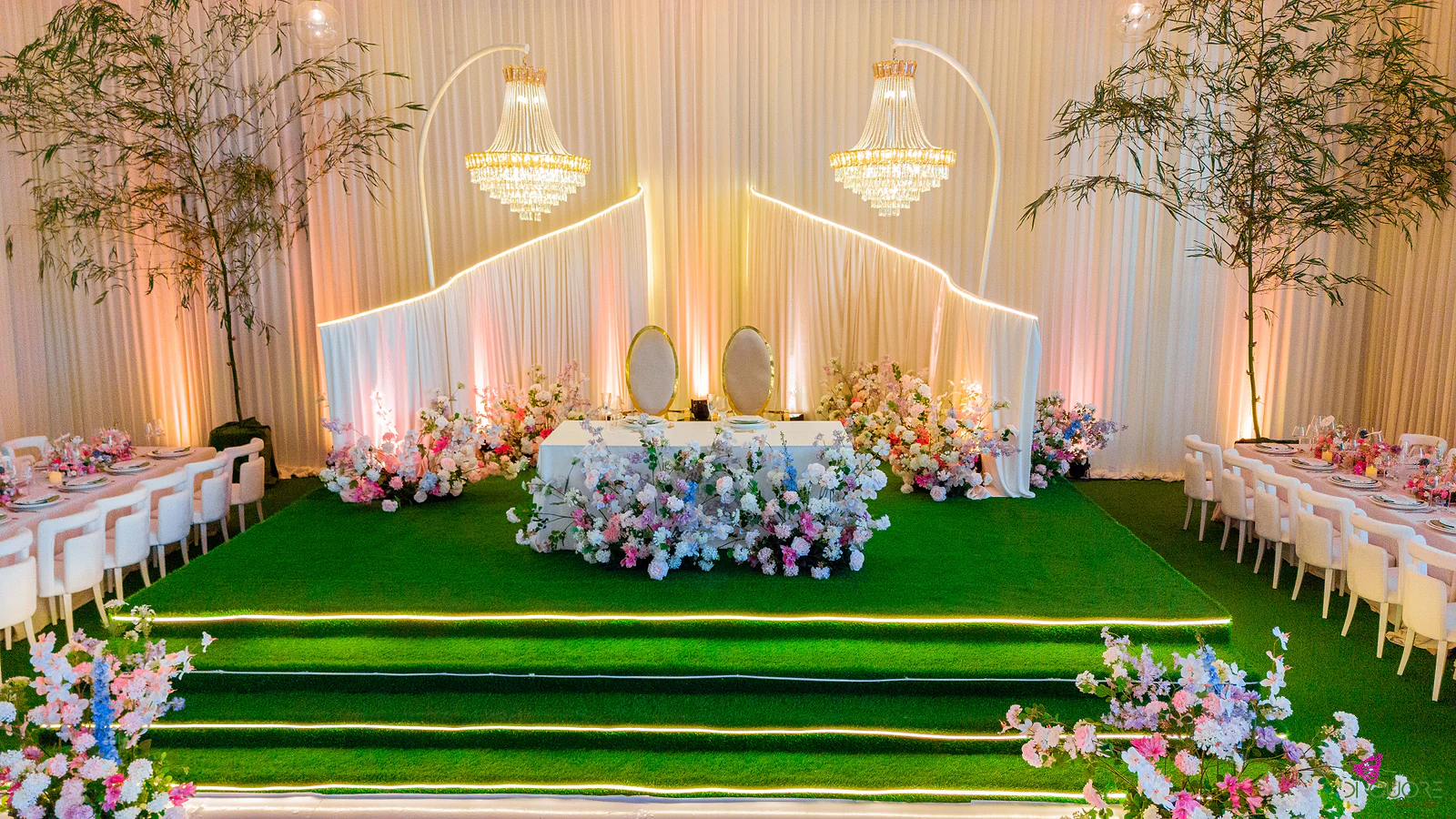 Mesa dos noivos sobre degraus forrados a verde com fita de luz, entre lustres e cortinados, com flores pastel na frente