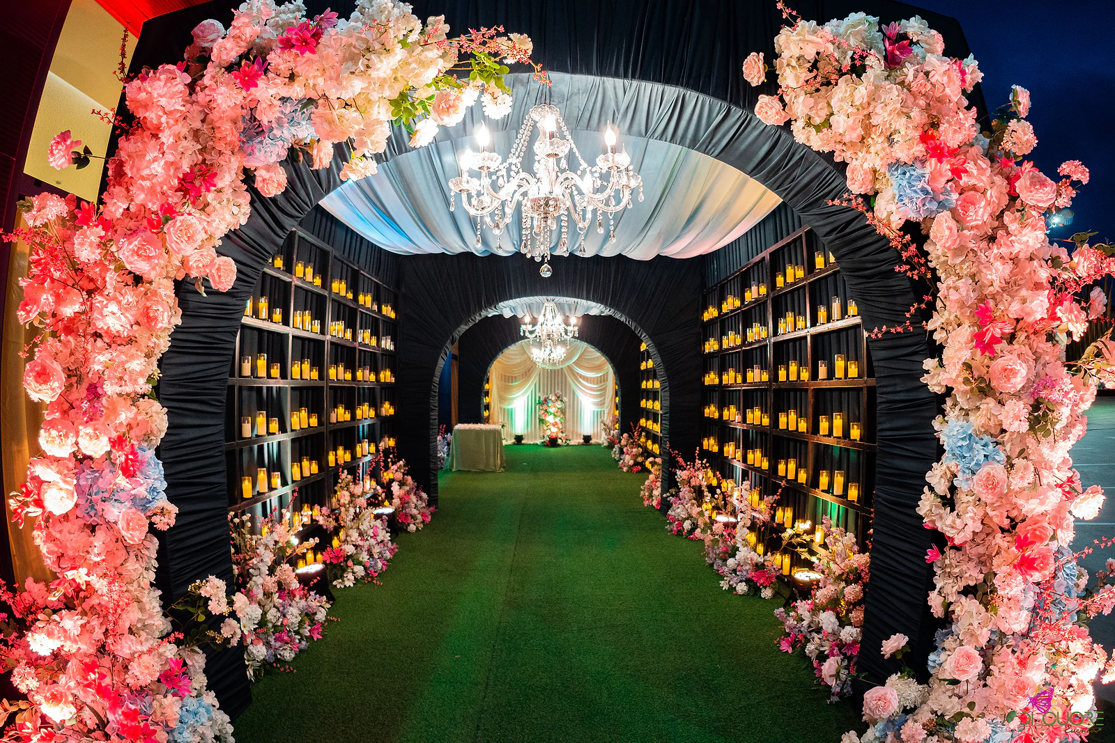Arco de flores cor-de-rosa e brancas à entrada de um túnel de tecido escuro com prateleiras de velas e um lustre, ao anoitecer