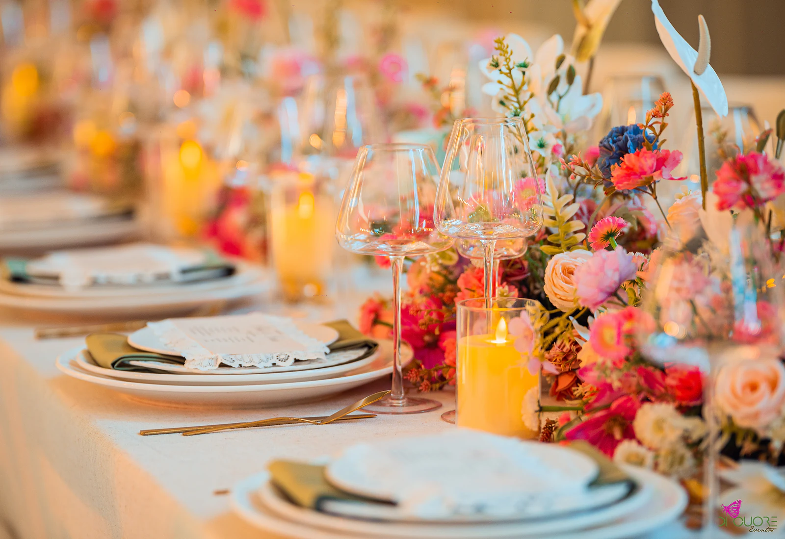 Mesa comprida posta com ramos baixos de flores cor-de-rosa e laranja, velas e copos de cristal