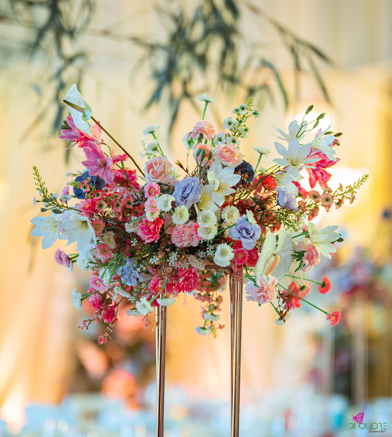 Arranjo alto de flores cor-de-rosa, lilás, brancas e vermelhas sobre um pedestal dourado, com luz quente e bambus desfocados ao fundo