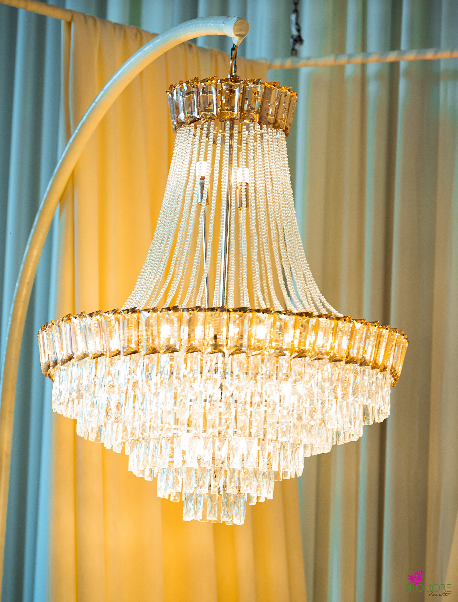 Lustre de cristal e dourado pendurado num arco metálico, diante de cortinados claros