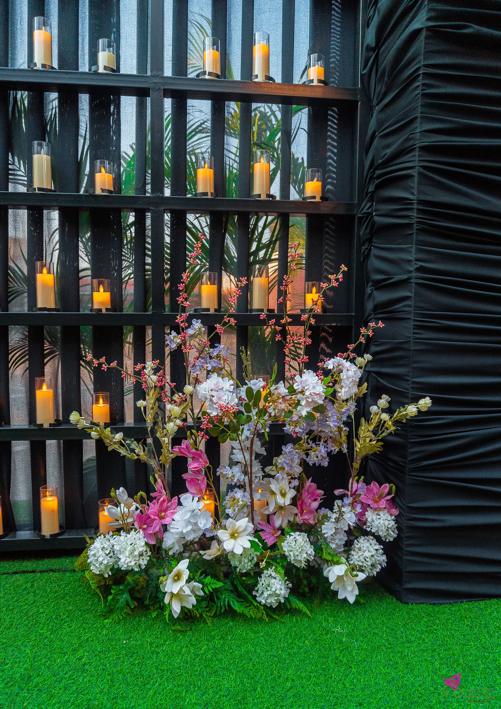 Maciço de flores brancas, cor-de-rosa e lilás junto a uma parede preta de prateleiras com velas acesas, sobre relva