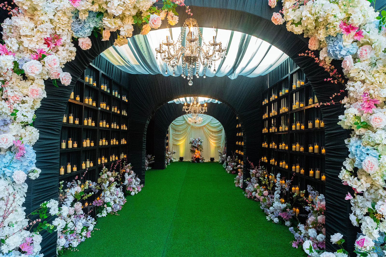Entrada do túnel de velas por um arco de flores brancas, cor-de-rosa e azuis, com prateleiras de velas, um lustre sob um tecto drapeado e relva ao centro