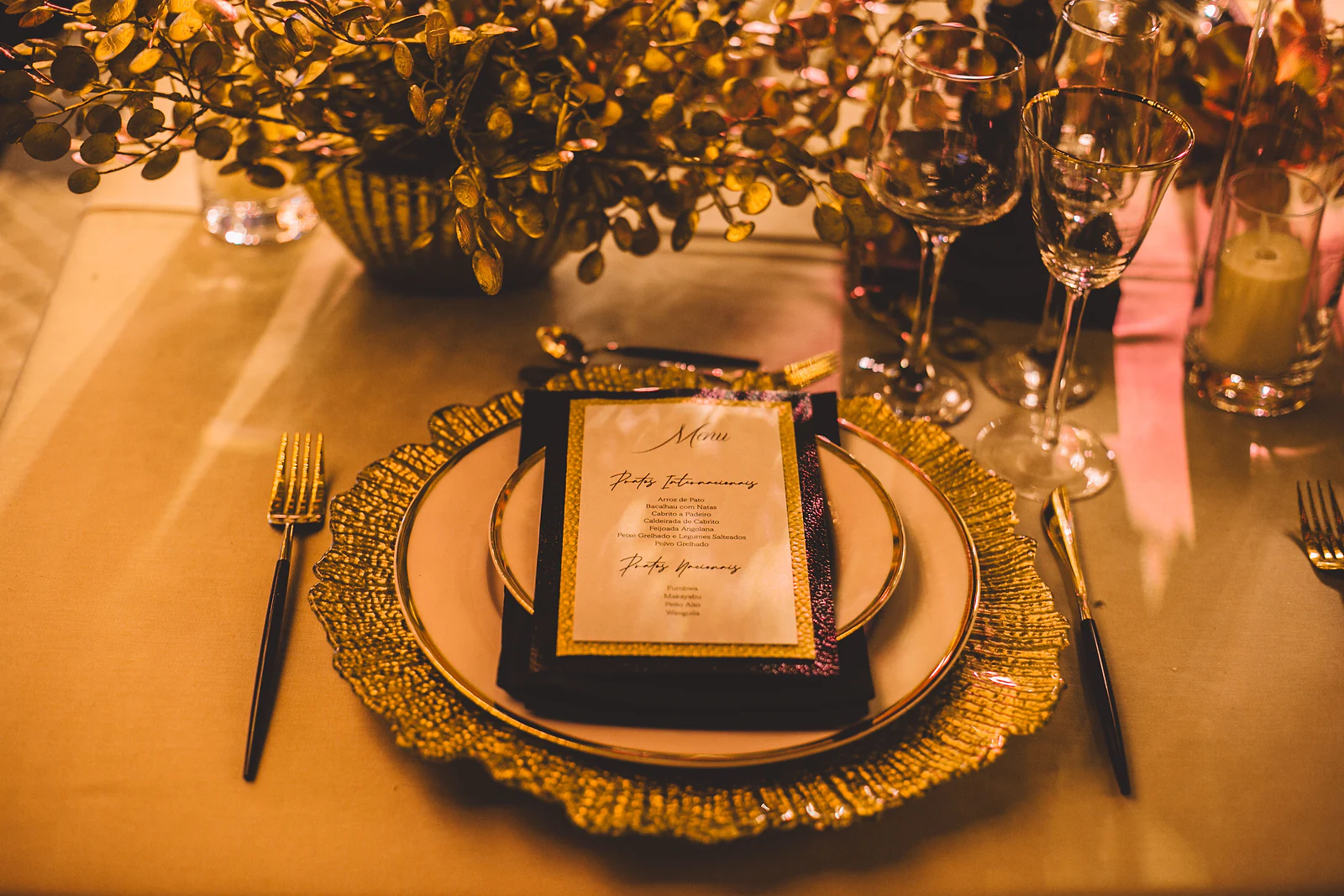 Prato de porcelana sobre sousplat dourado com menu impresso, entre talheres de cabo preto e um arranjo de folhas douradas