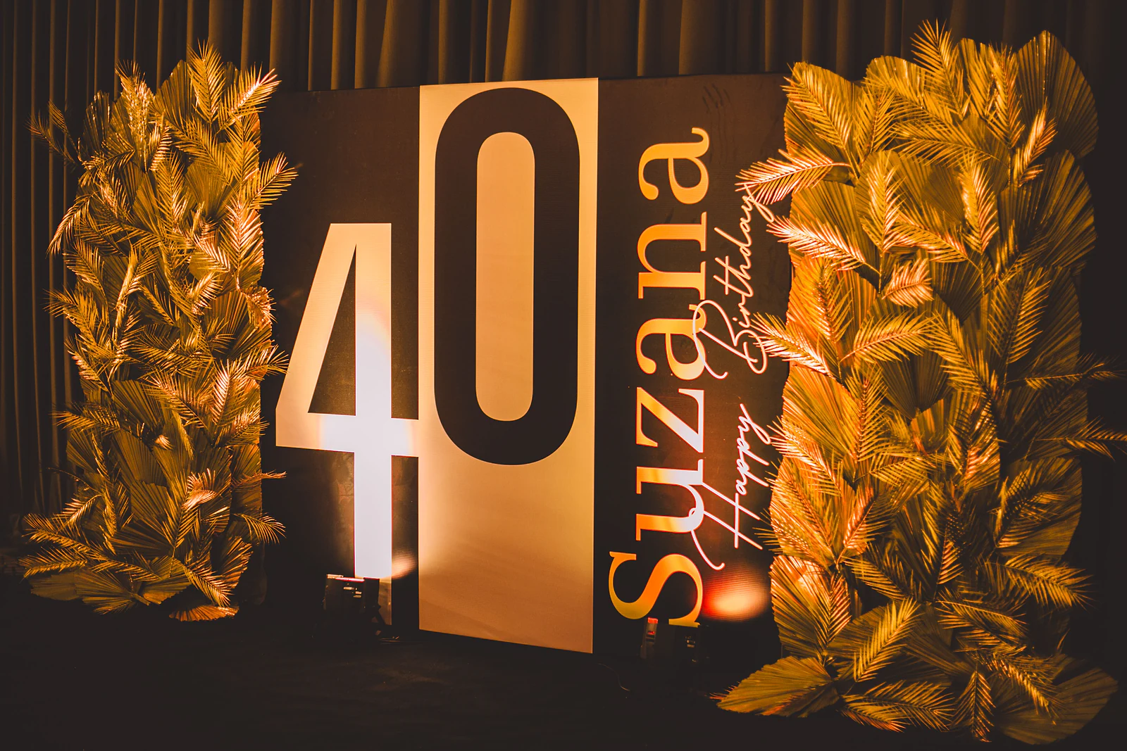 Letreiro «40 Suzana Happy Birthday» entre dois painéis de palmas douradas, diante de um cortinado escuro