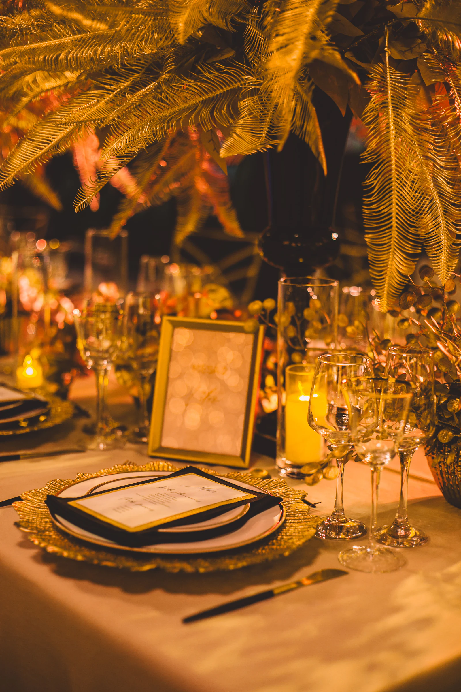Moldura dourada com número de mesa, menu sobre prato com sousplat dourado e palmas douradas por cima, com velas ao lado