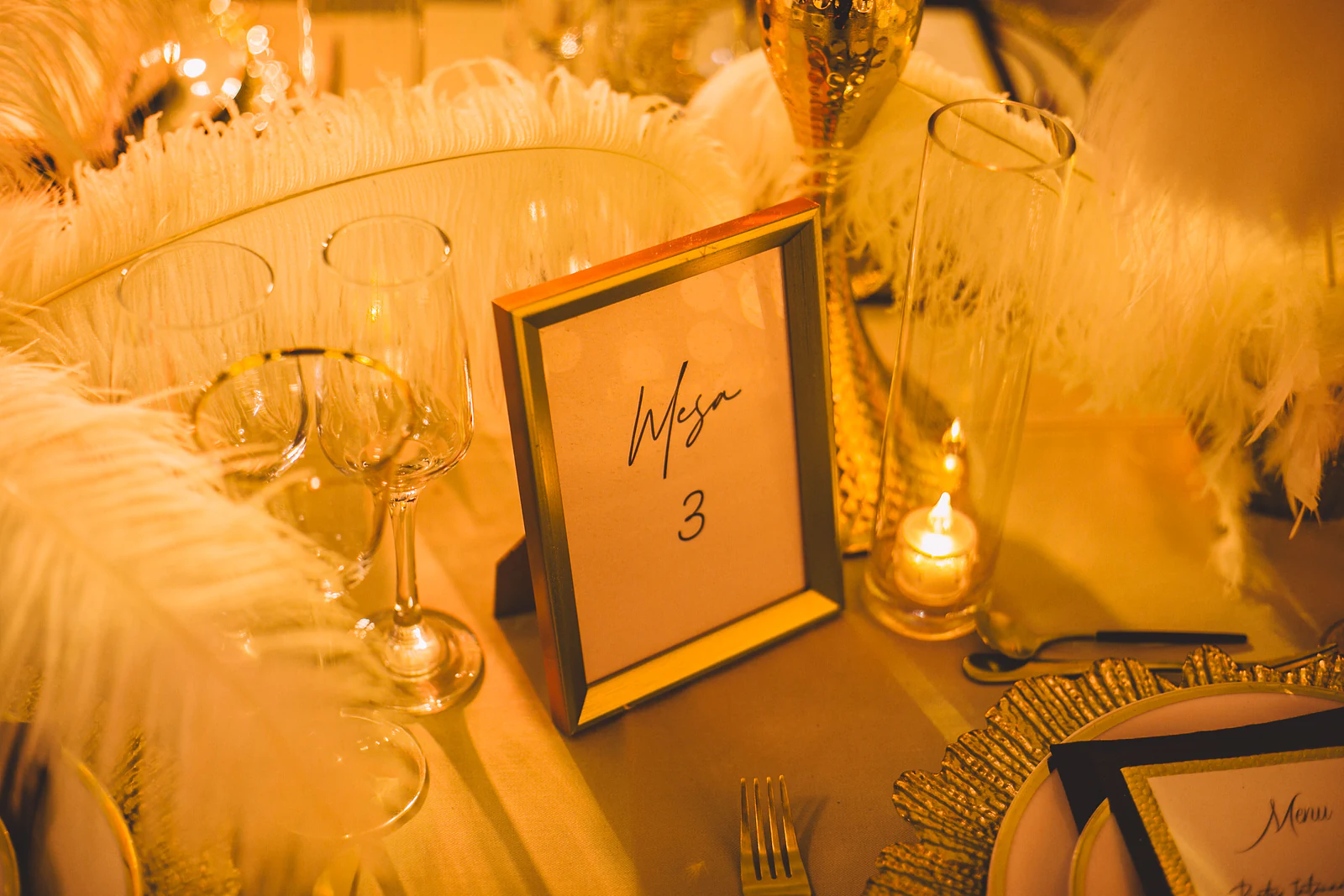 Moldura dourada com «Mesa 3» entre plumas brancas, copos e uma vela, junto a um sousplat dourado com menu