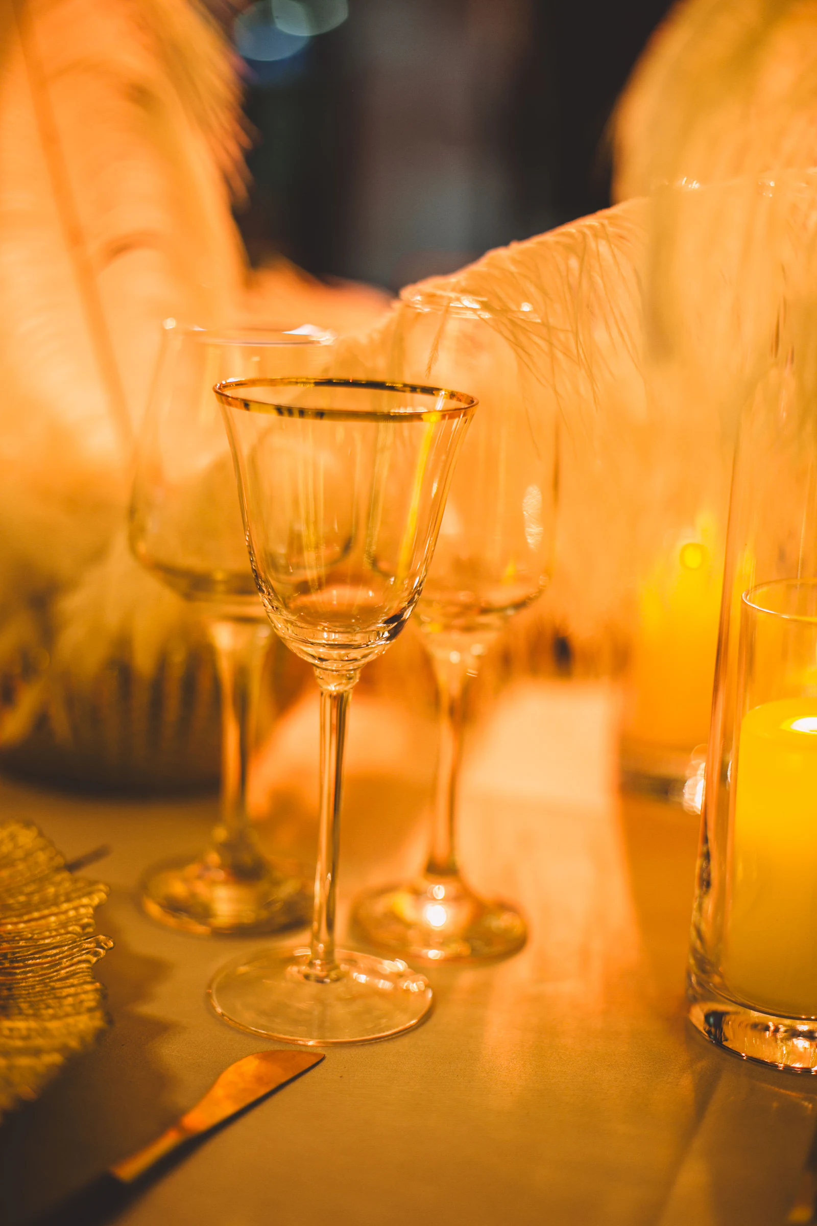 Taças de rebordo dourado, velas e plumas brancas sobre a mesa, em luz quente