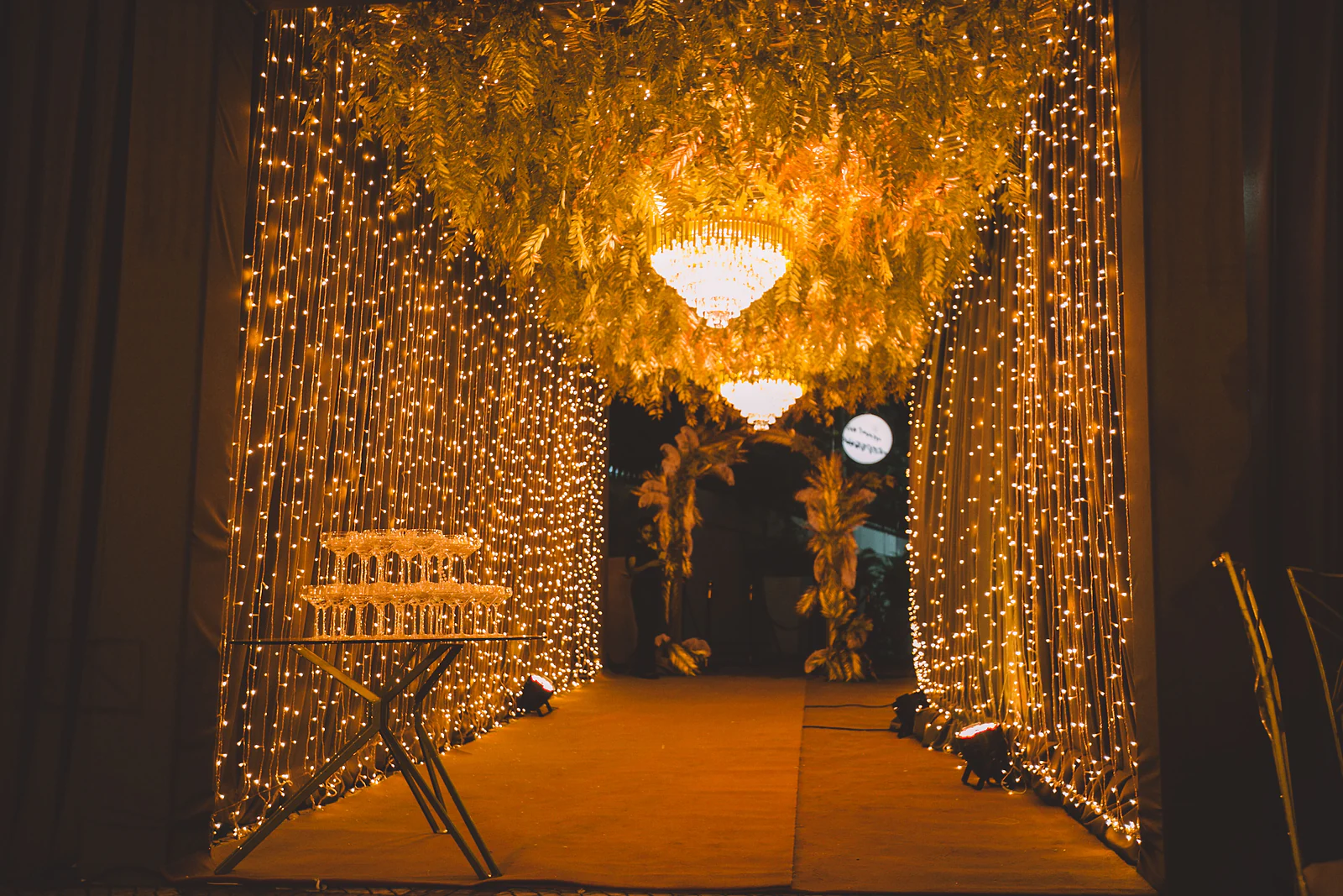 Túnel de fios de luz com tecto de folhagem dourada e candeeiros de cristal, com uma torre de taças de champanhe à esquerda
