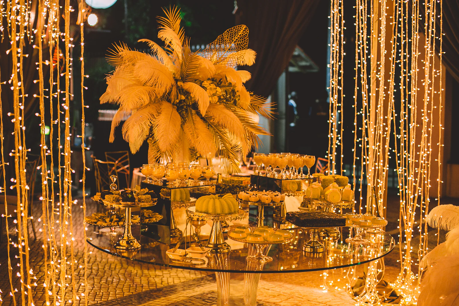 Mesa de vidro redonda com doces e taças sobre bases douradas e um grande arranjo de plumas, entre cortinas de fios de luz