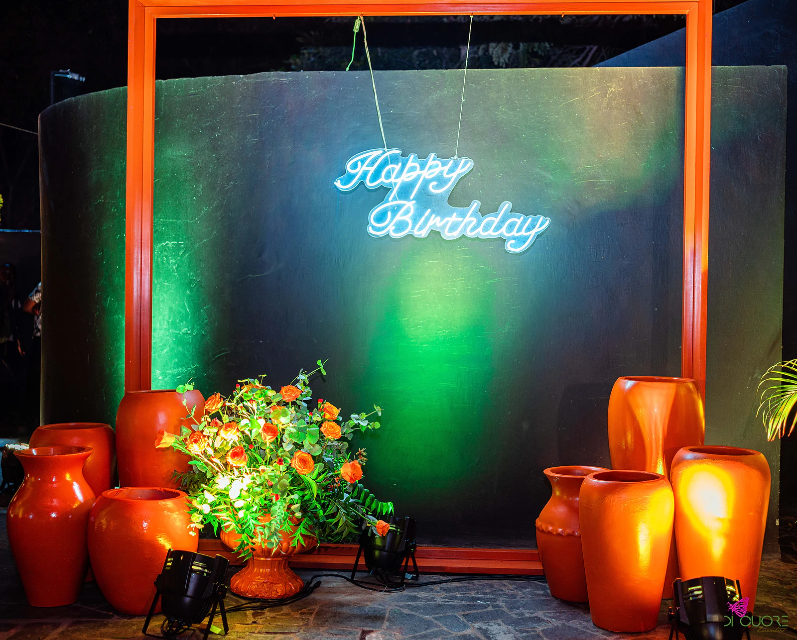 Vista frontal da moldura laranja com o néon “Happy Birthday” sobre o painel verde-escuro, flanqueada por jarros de barro, com um arranjo de rosas laranja à esquerda e projectores no chão
