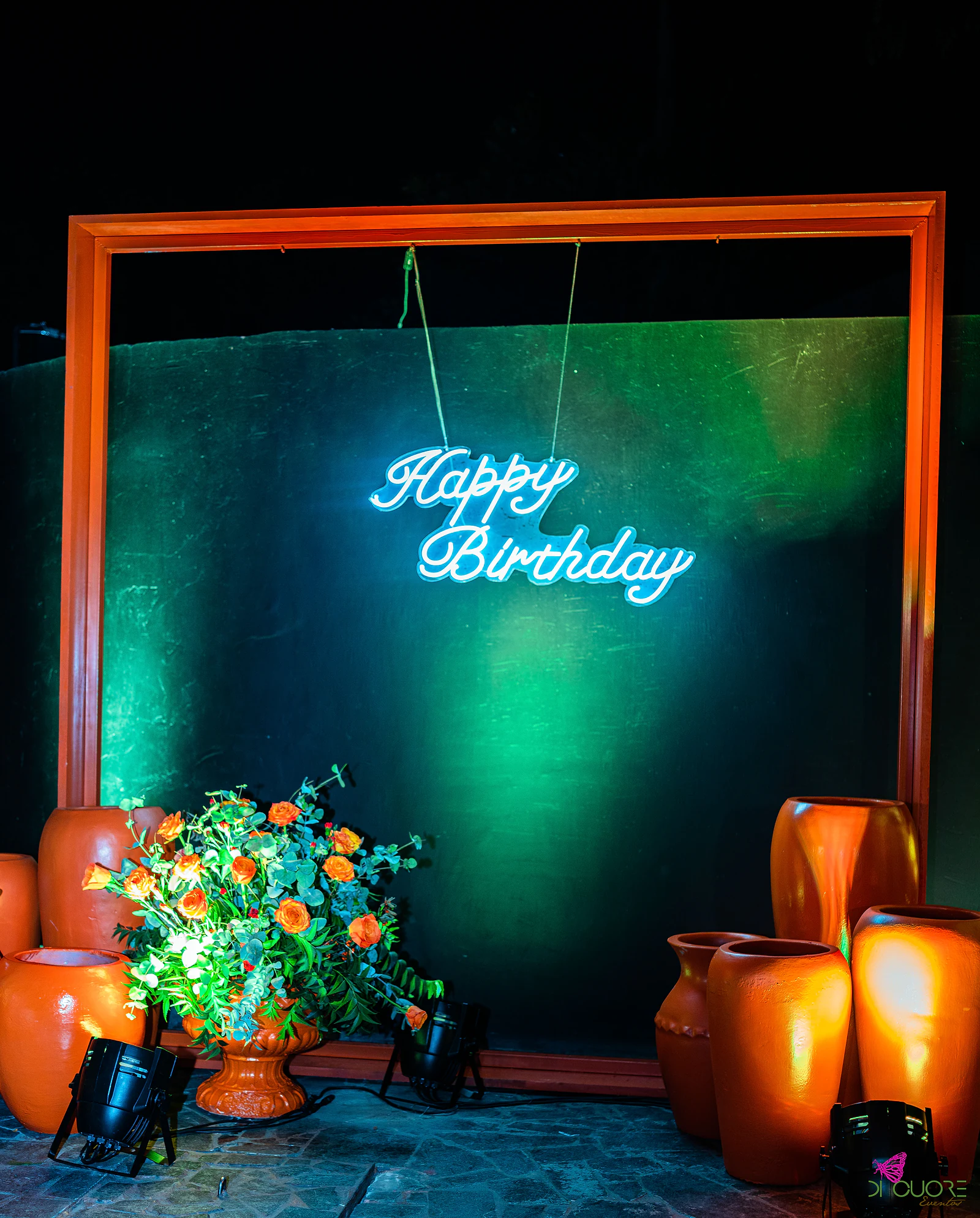 Moldura laranja com letreiro de néon “Happy Birthday” sobre um painel verde-escuro, rodeada de jarros de barro e de um arranjo de rosas laranja