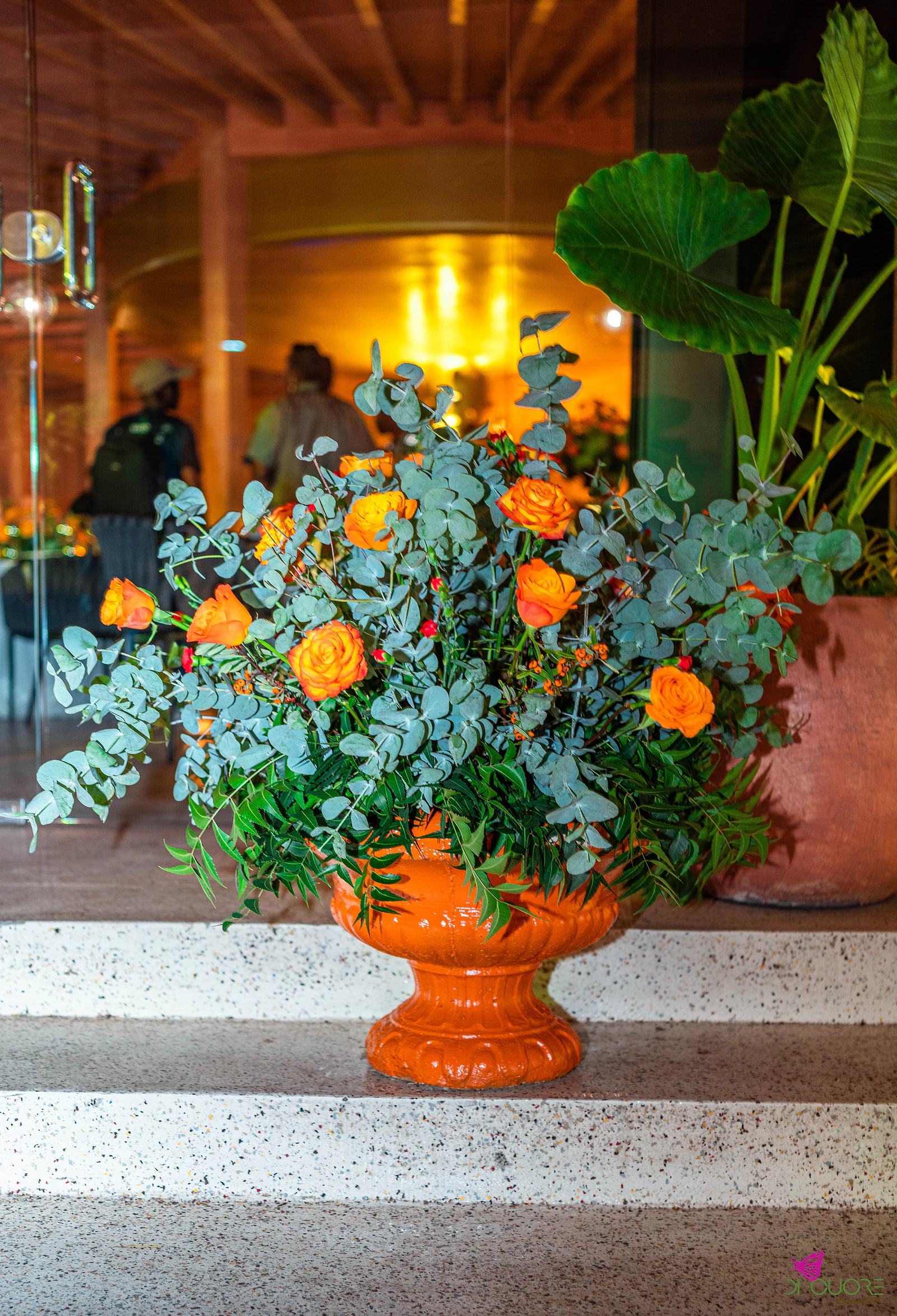 Arranjo de rosas laranja, botões vermelhos e eucalipto numa urna laranja sobre degraus de pedra clara, junto a uma planta de folhas largas, com o salão iluminado em âmbar ao fundo