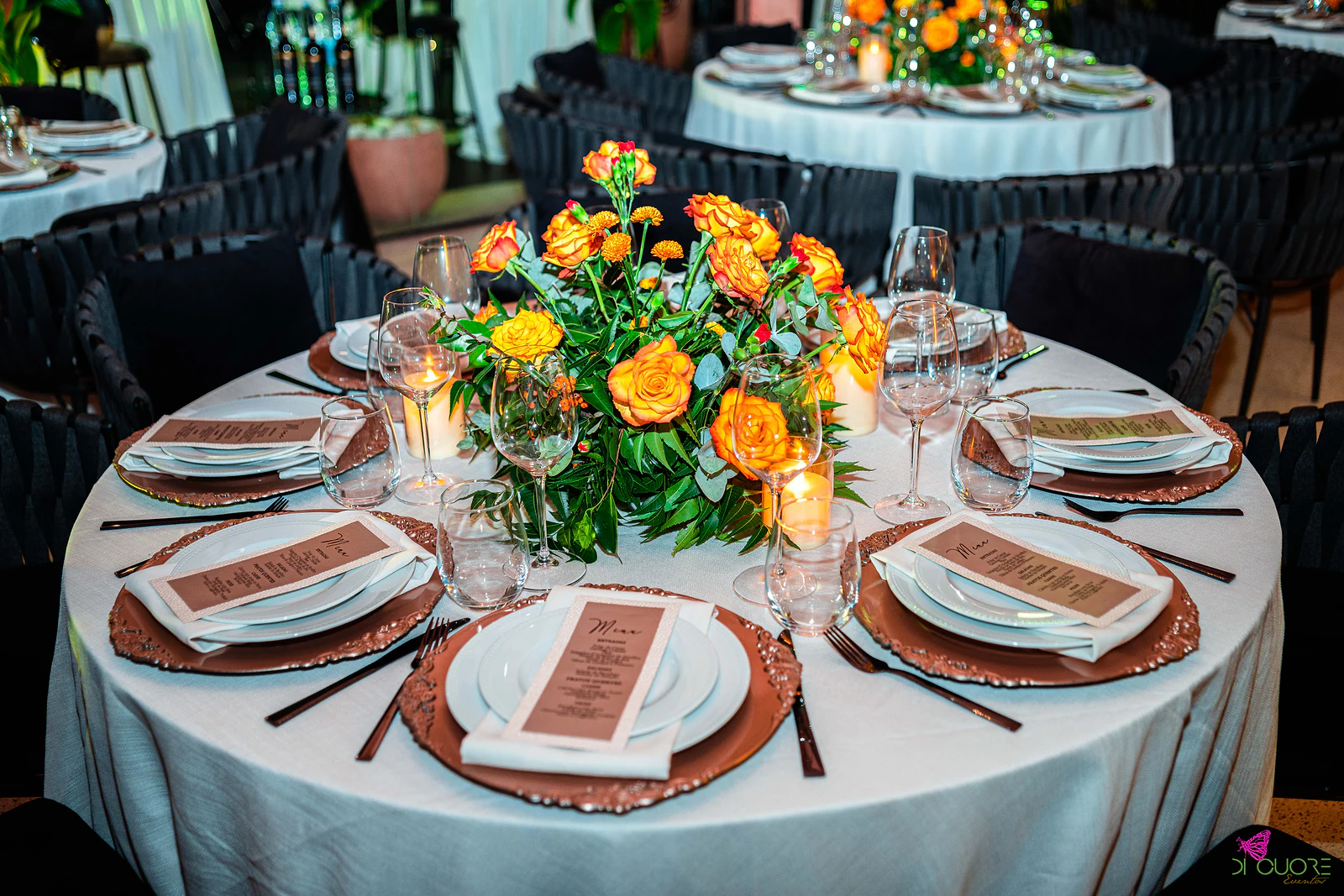 Mesa redonda de toalha branca posta com sousplats cobre, menus impressos e velas, com um centro de rosas e cravos laranja e outras mesas ao fundo