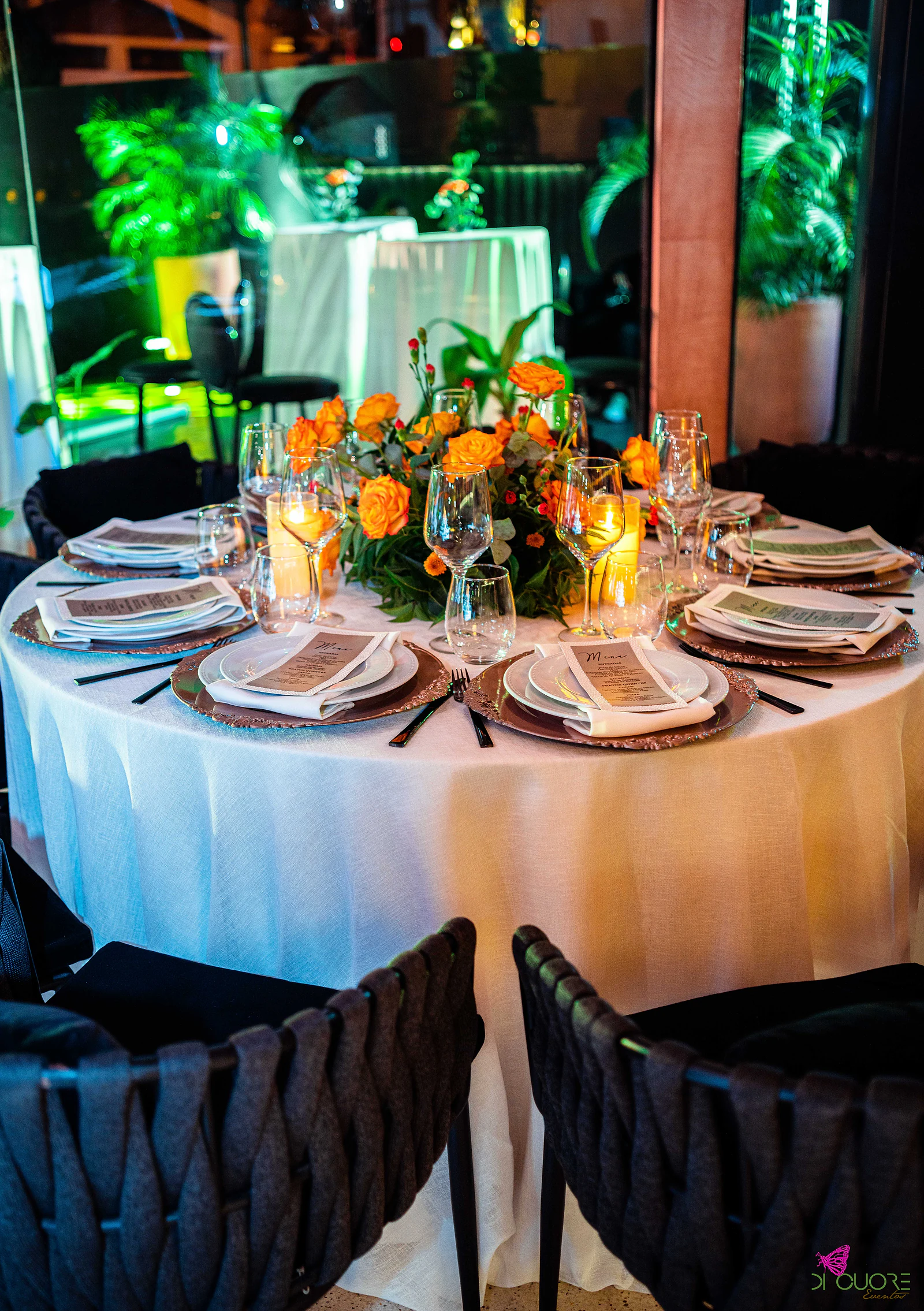 Mesa redonda de toalha clara com sousplats cobre, menus, velas e centro de rosas laranja, entre cadeiras pretas de corda entrançada
