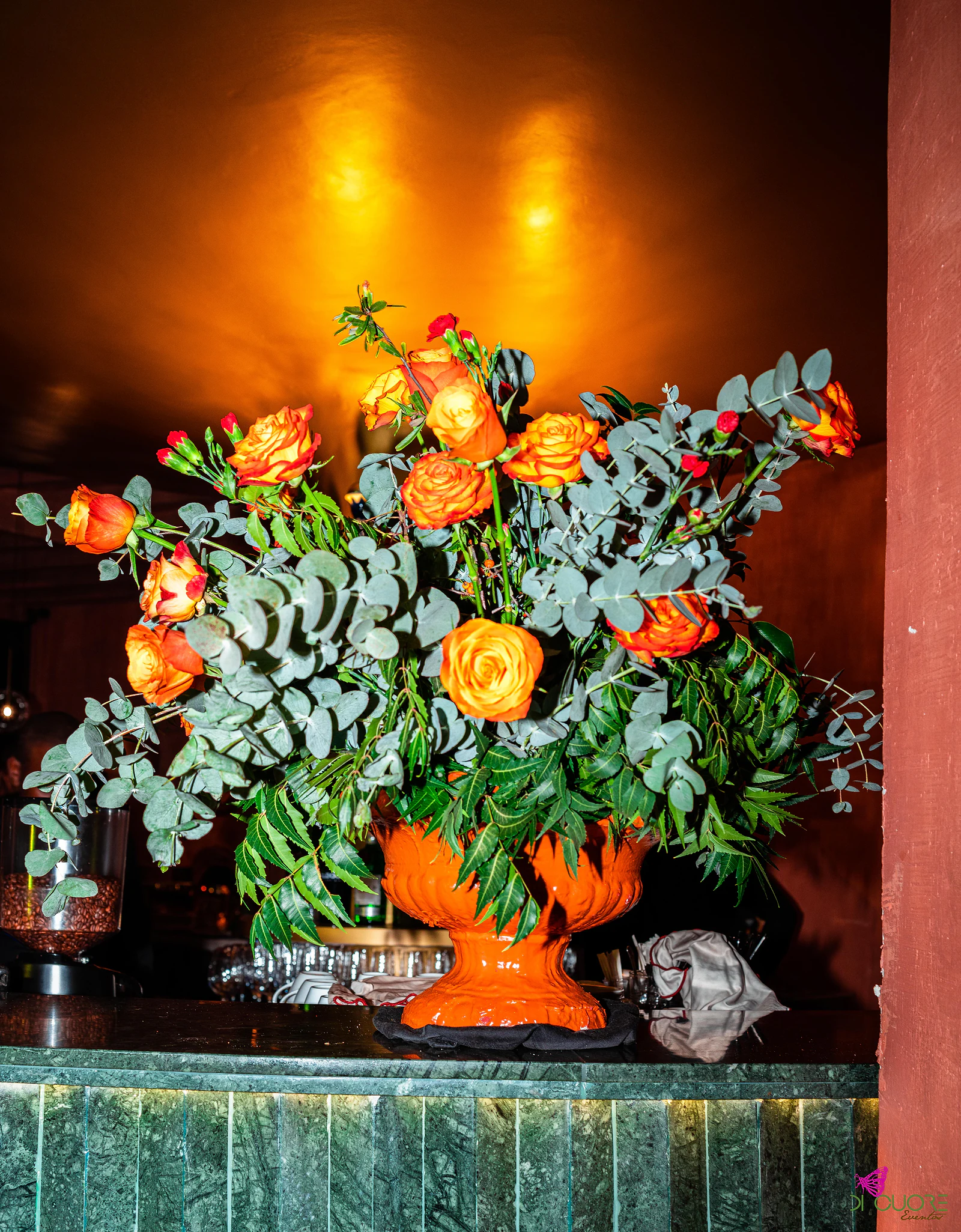 Grande arranjo de rosas laranja e eucalipto numa urna laranja sobre um balcão de mármore verde, sob luz dourada