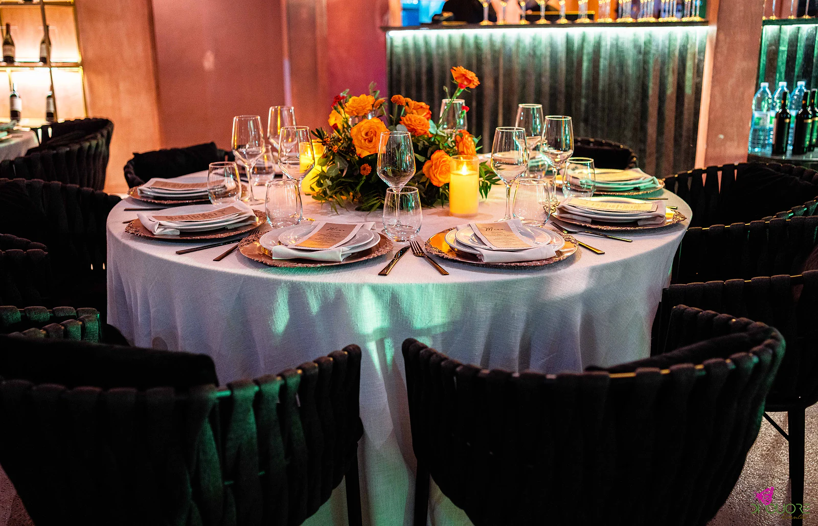 Mesa redonda posta com sousplats cobre e centro de rosas laranja, diante de um balcão de mármore verde iluminado, com prateleiras de garrafas de um dos lados