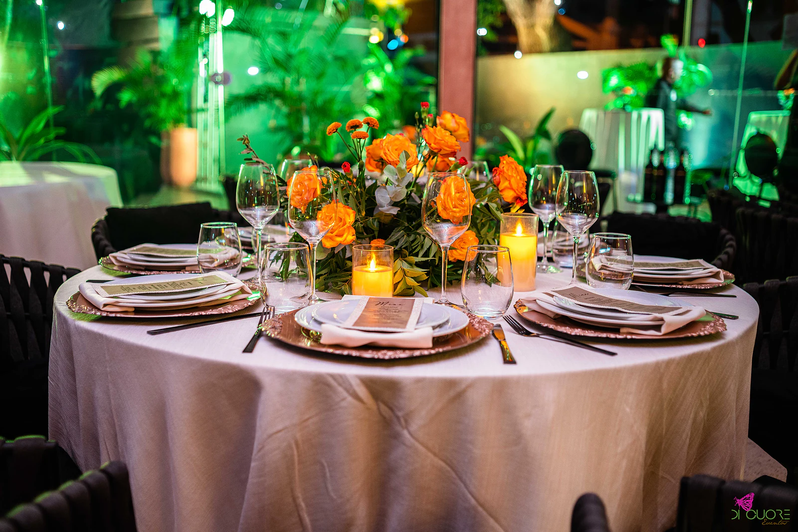 Mesa redonda de toalha clara posta com sousplats cobre e menus, velas e centro de rosas laranja, com folhagem iluminada a verde ao fundo