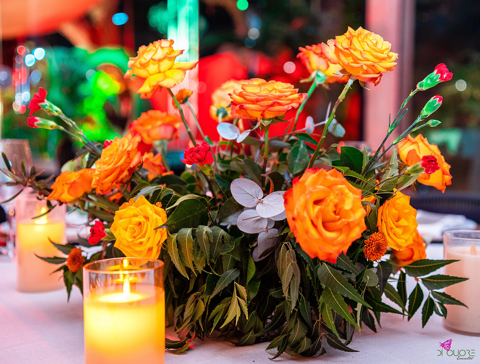 Rosas laranja e cravos vermelhos com eucalipto, ao lado de velas em copos de vidro