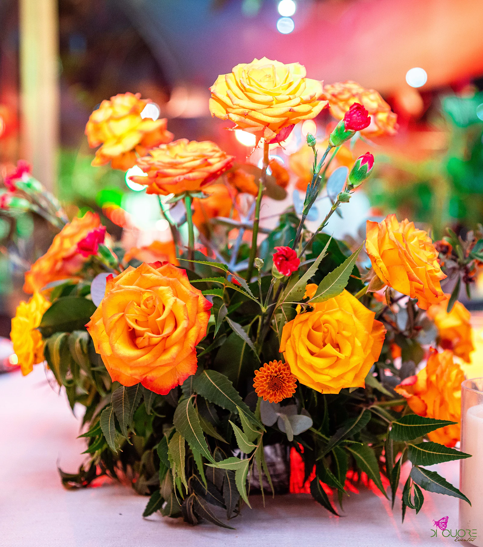 Rosas laranja e amarelas com cravos vermelhos e folhagem num centro de mesa, com uma vela em copo de vidro ao canto e luzes desfocadas ao fundo