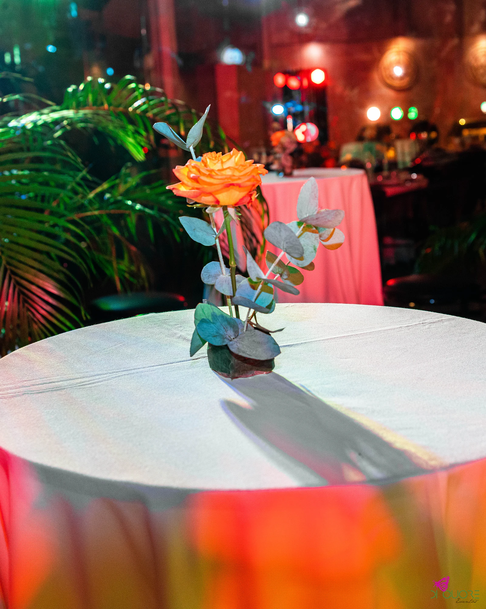 Uma rosa laranja com ramos de eucalipto num pequeno vaso sobre uma mesa alta de toalha clara, com palmeiras e uma segunda mesa cor-de-rosa ao fundo