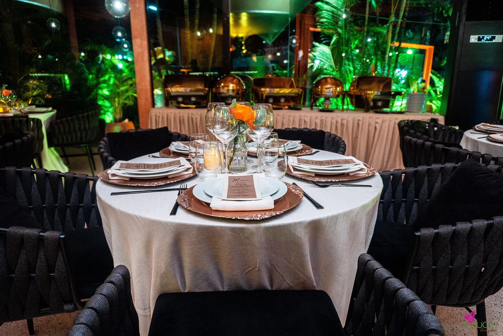 Mesa redonda posta com sousplats cobre, menus e velas, com uma rosa laranja numa jarra de vidro, diante do buffet de recipientes de cobre e de palmeiras iluminadas a verde